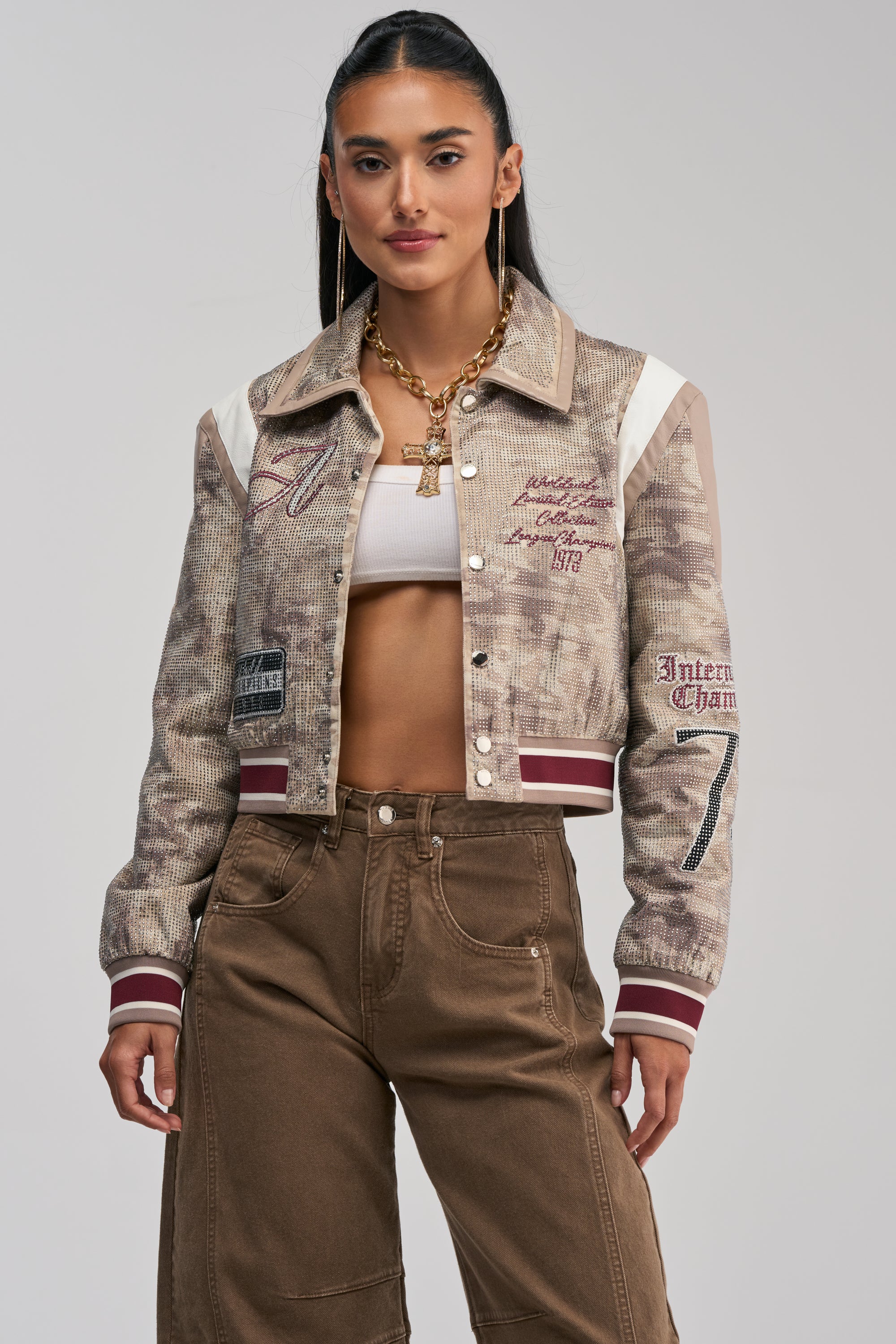 A woman with long dark hair stands against a plain light background, showcasing statement street style in the BIG EGO RHINESTONE BOMBER IN CAMO, layered over a white crop top and brown pants and accessorized with gold jewelry.