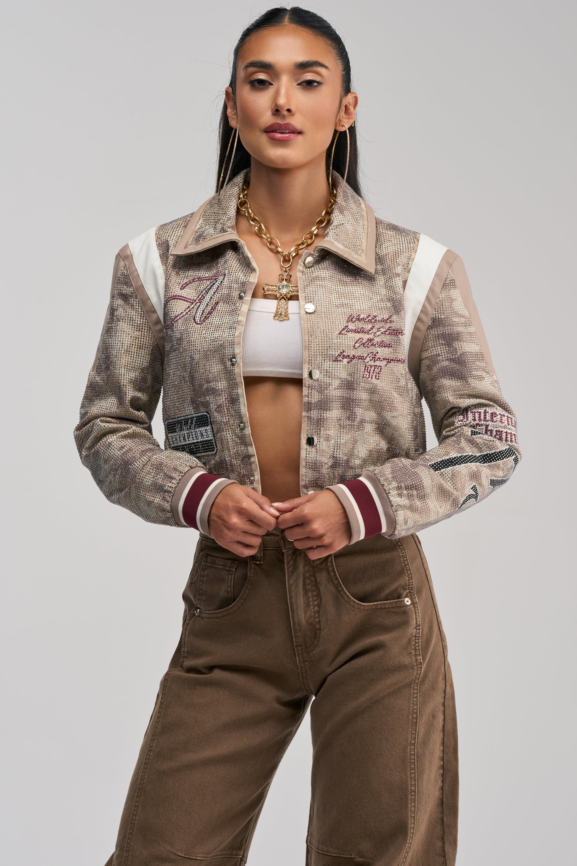 A woman with long dark hair, gold jewelry, and hoop earrings exudes statement street style as she poses confidently against a plain background in the BIG EGO RHINESTONE BOMBER IN CAMO, styled with a white crop top and high-waisted brown pants.
