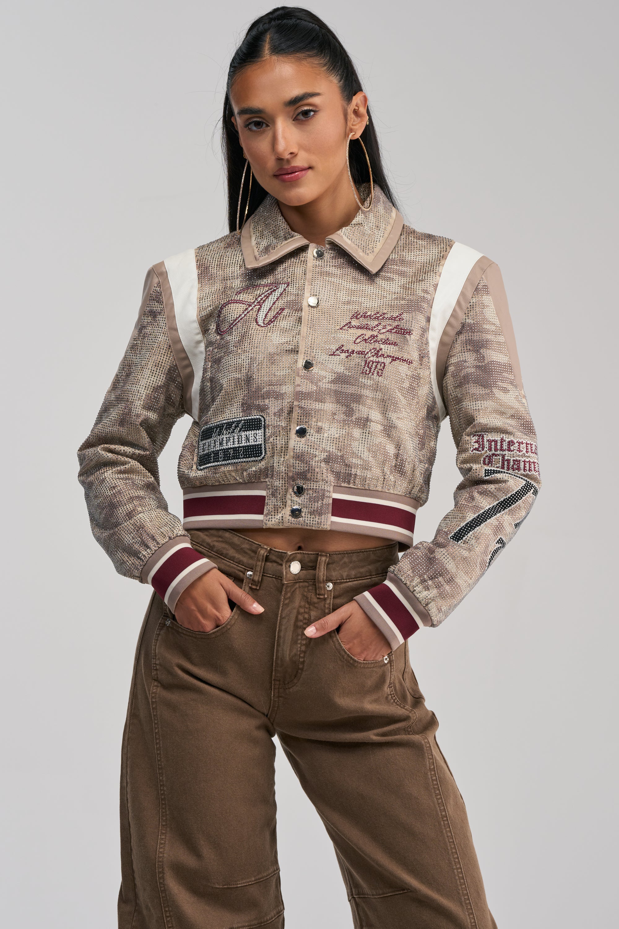 Wearing the BIG EGO RHINESTONE BOMBER IN CAMO, styled with brown high-waisted pants, she showcases statement street style as she poses with her hands in her pockets against a plain light background.