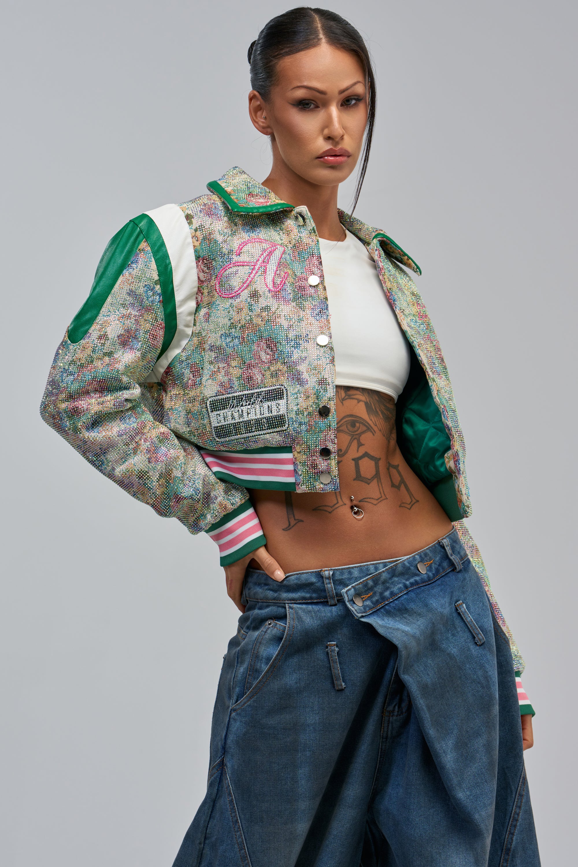A woman with long dark hair in braided pigtails and tattoos poses seriously in the TAPPED OUT RHINESTONE BOMBER, paired with a white crop top and baggy jeans.