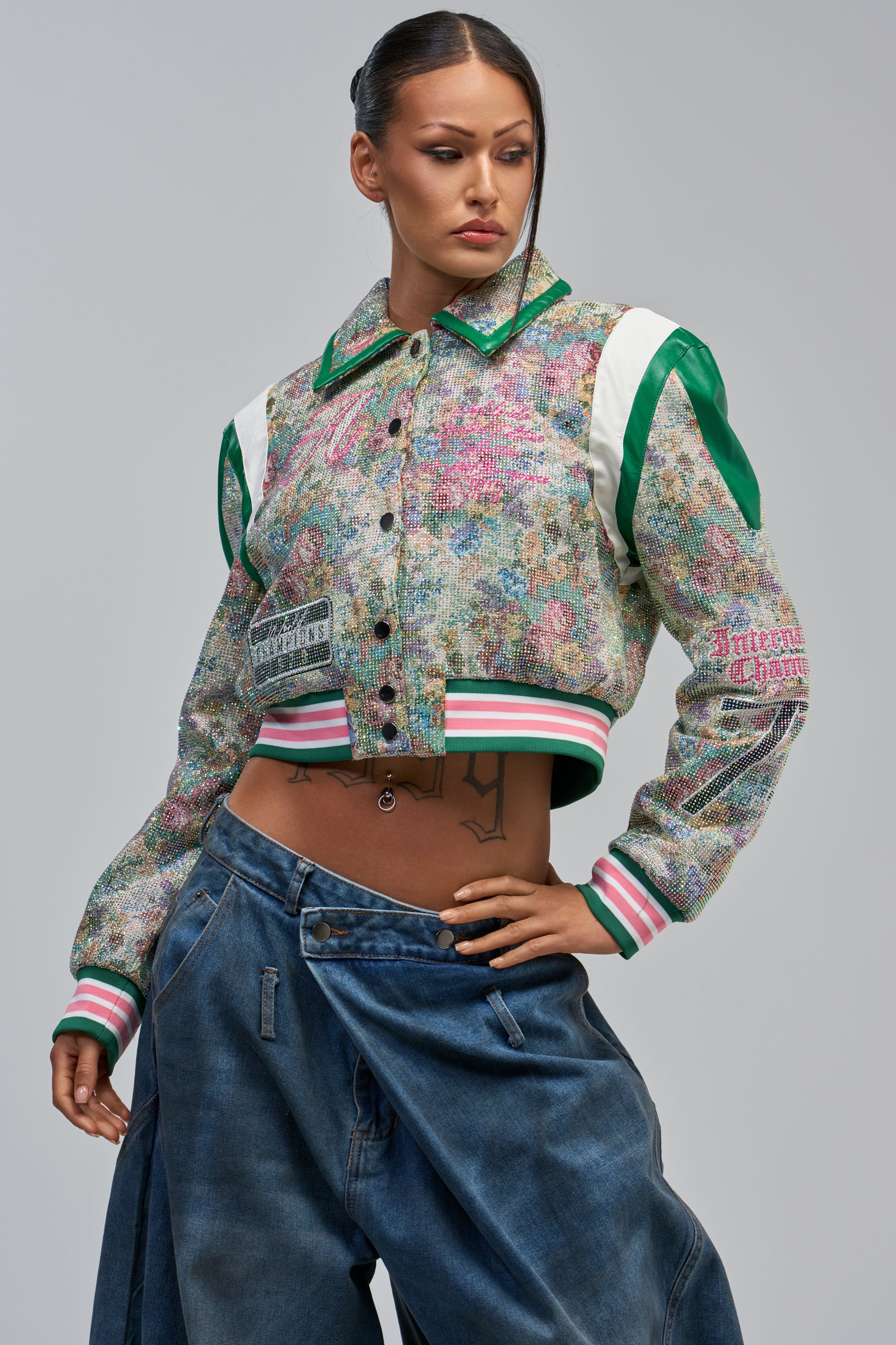 Wearing the TAPPED OUT RHINESTONE BOMBER with green and pink accents and loose blue jeans, a person poses confidently with one hand on their hip against a plain background.