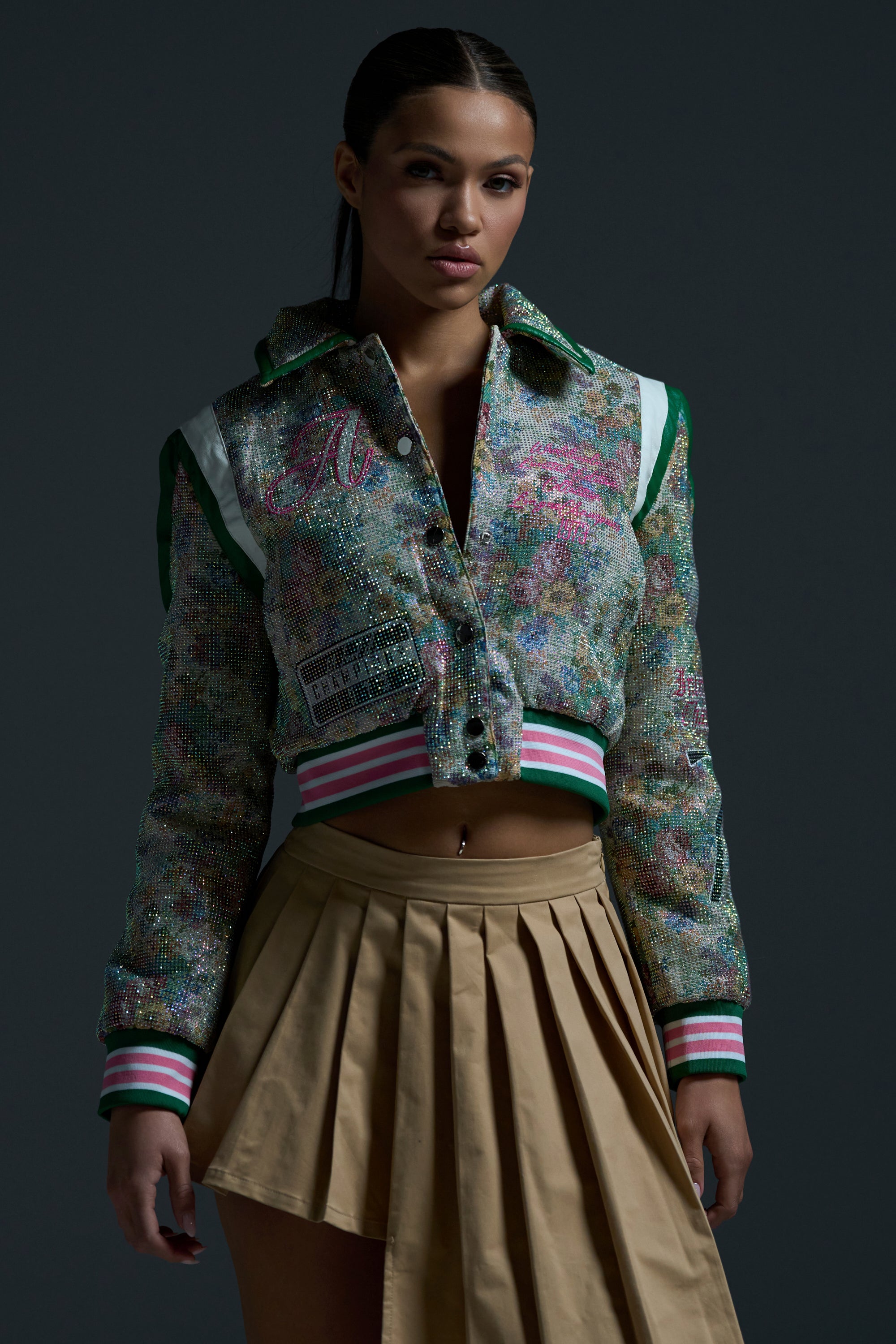 A woman stands against a dark background wearing the TAPPED OUT RHINESTONE BOMBER with striped cuffs and faux leather trim, paired with a beige pleated skirt. Her slicked-back dark hair and confident expression complete the look.