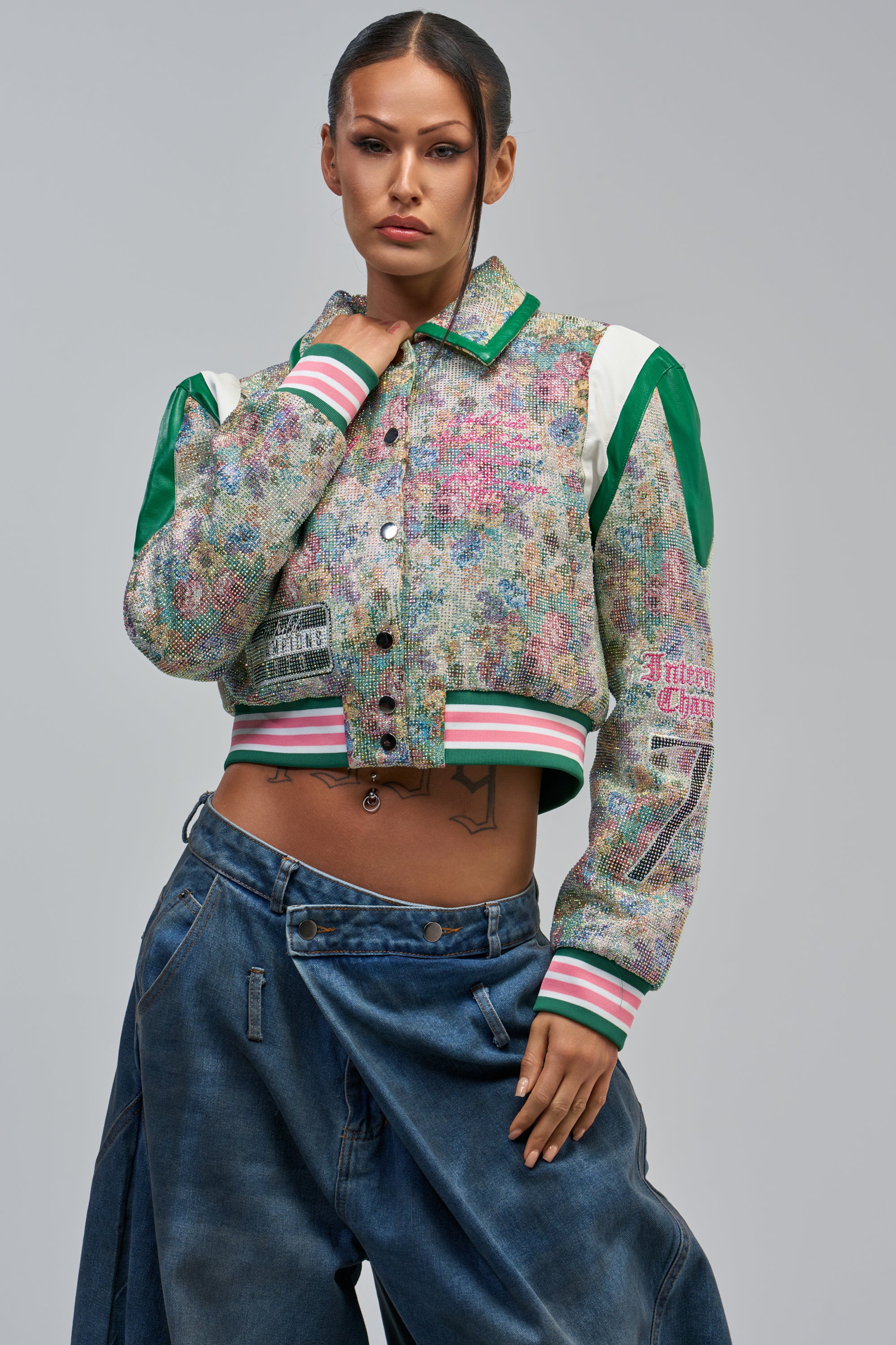 A woman showcases modern streetwear in the TAPPED OUT RHINESTONE BOMBER paired with loose-fitting jeans, posing confidently against a plain gray background.