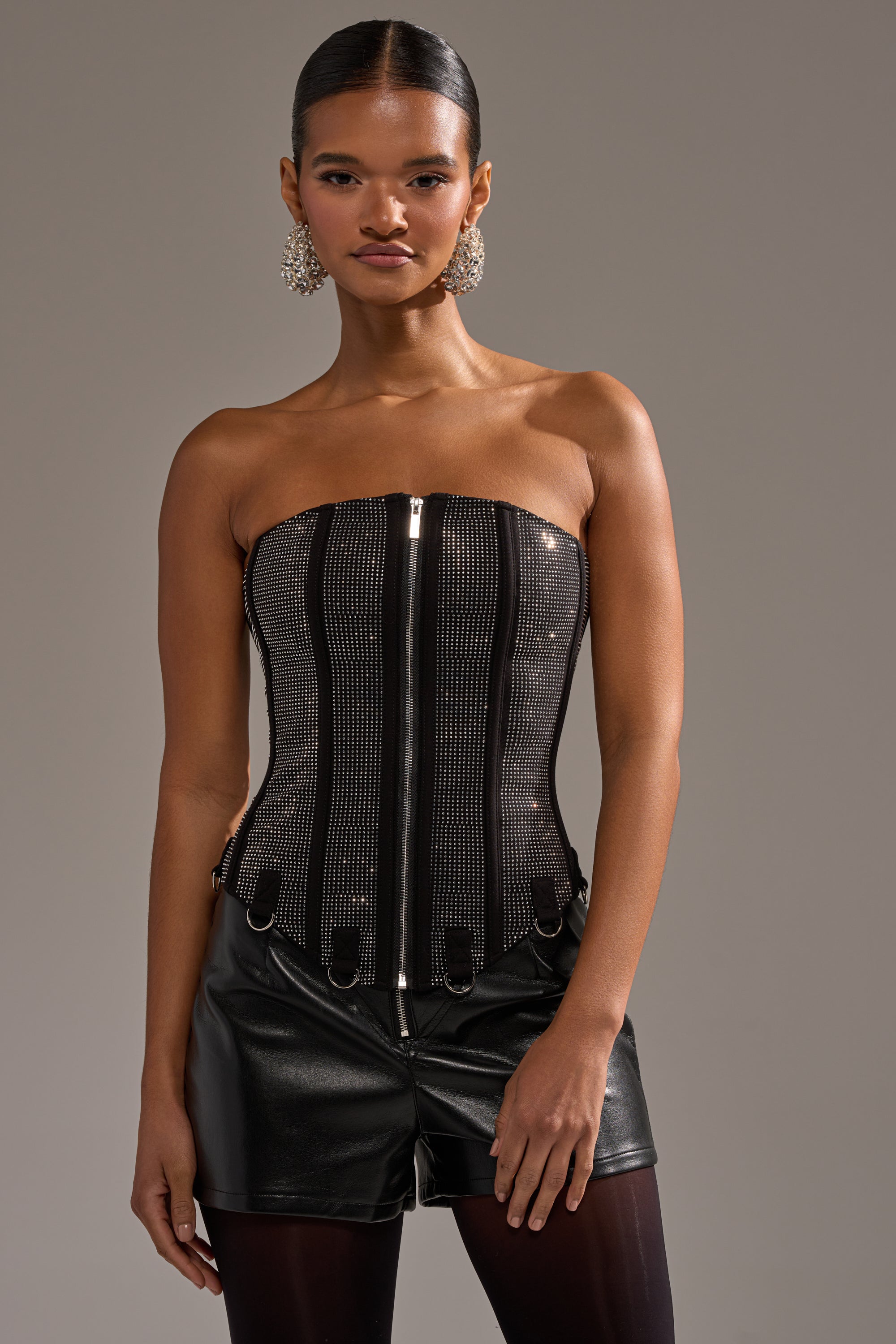 A woman models the MORE CHAMPAGNE PLEASE RHINESTONE CORSET with silver studs, paired with black leather shorts, black tights, and statement earrings against a plain gray background.