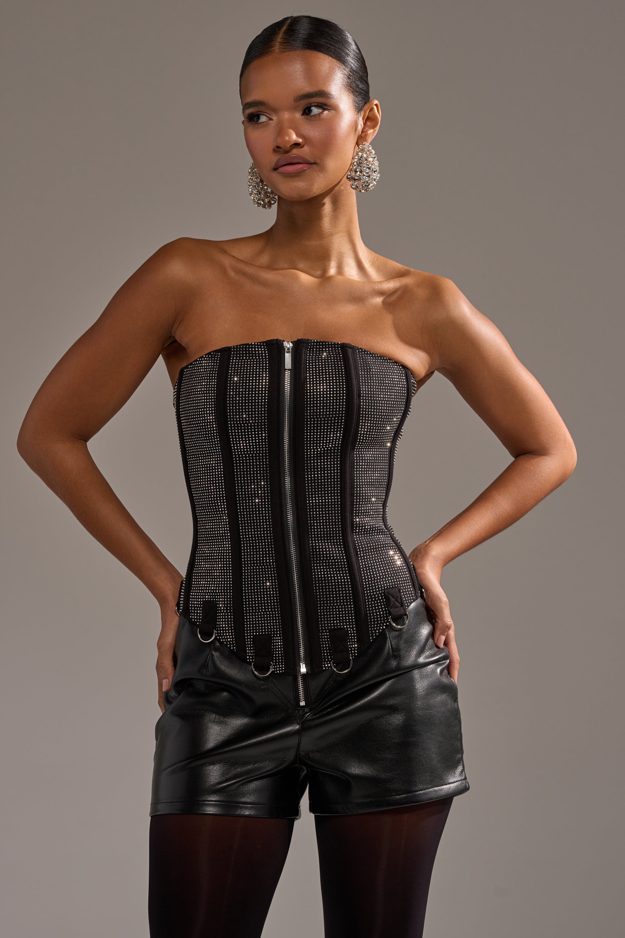 A woman poses with hands on hips in the MORE CHAMPAGNE PLEASE RHINESTONE CORSET, styled with black leather shorts, black tights, and bold statement earrings against a neutral background.