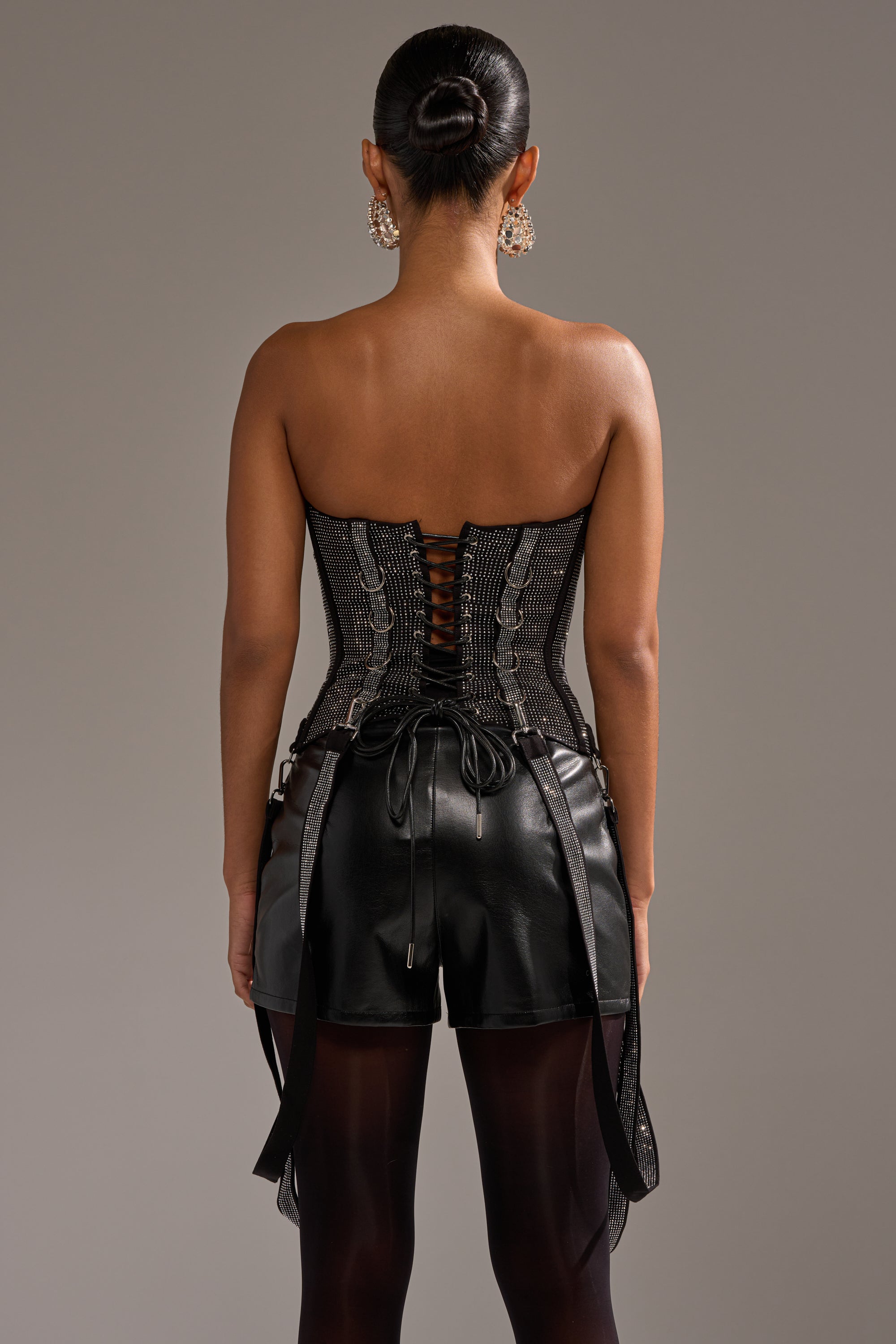 A person faces away in the MORE CHAMPAGNE PLEASE RHINESTONE CORSET, black leather shorts with side ties, sheer black tights, large earrings, and a sleek bun, set against a plain gray background.