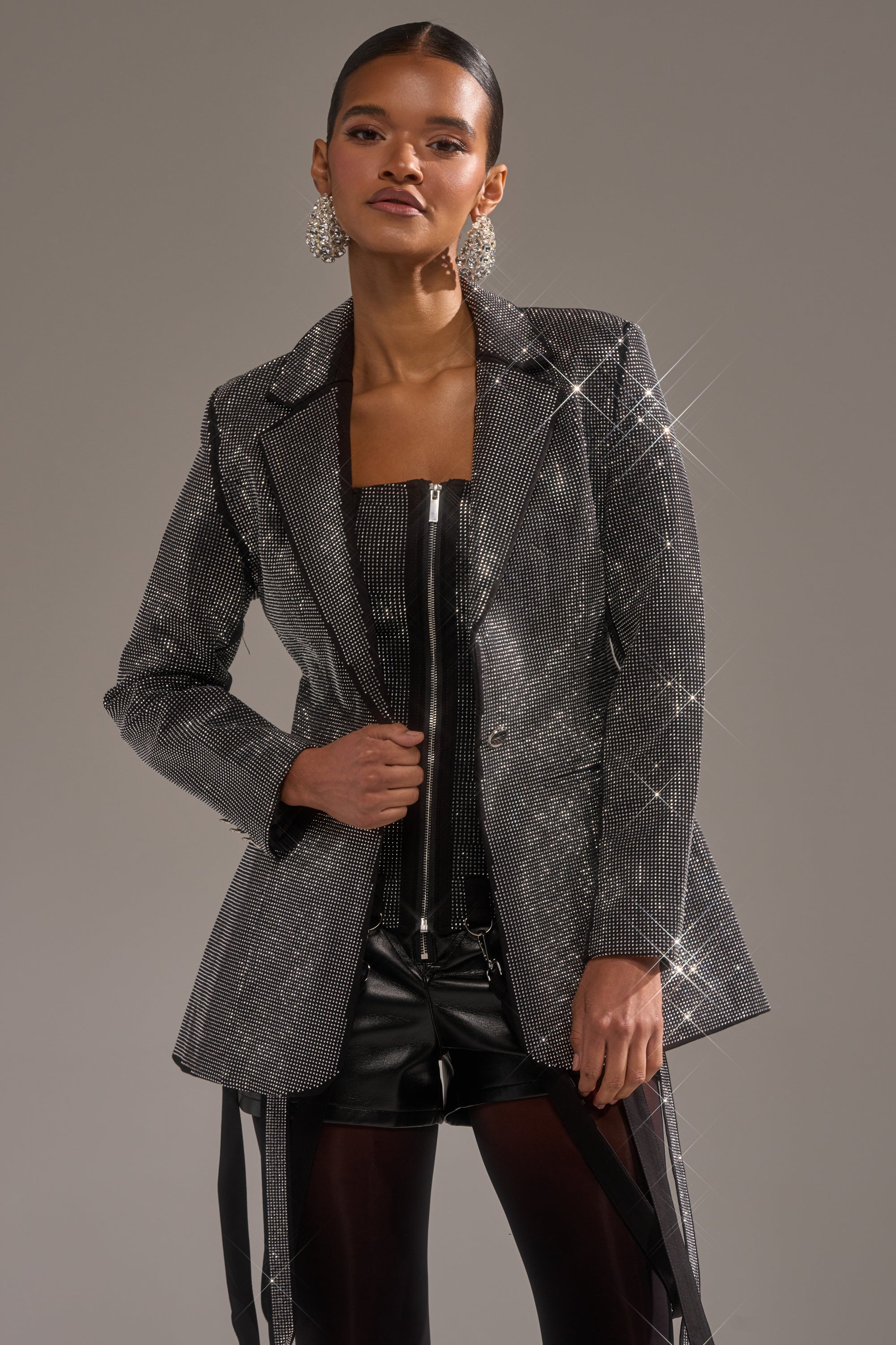 A woman poses confidently in the "MORE CHAMPAGNE PLEASE BACKLESS RHINESTONE BLAZER" over a black zippered dress and shorts, styled with sheer tights and statement earrings against a plain background.