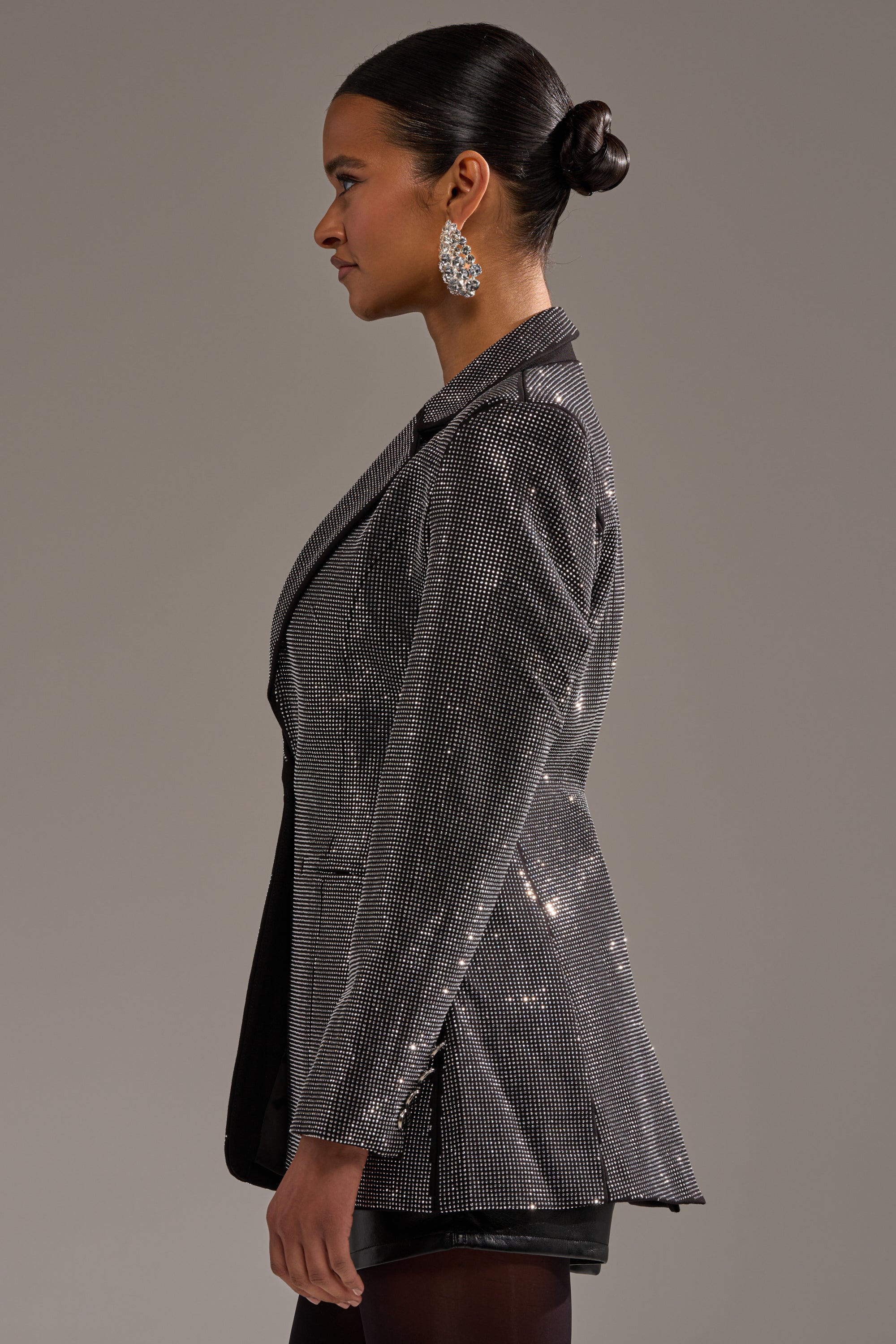 A woman in profile with a sleek bun and statement earrings wears the MORE CHAMPAGNE PLEASE BACKLESS RHINESTONE BLAZER over black, standing against a plain gray background.