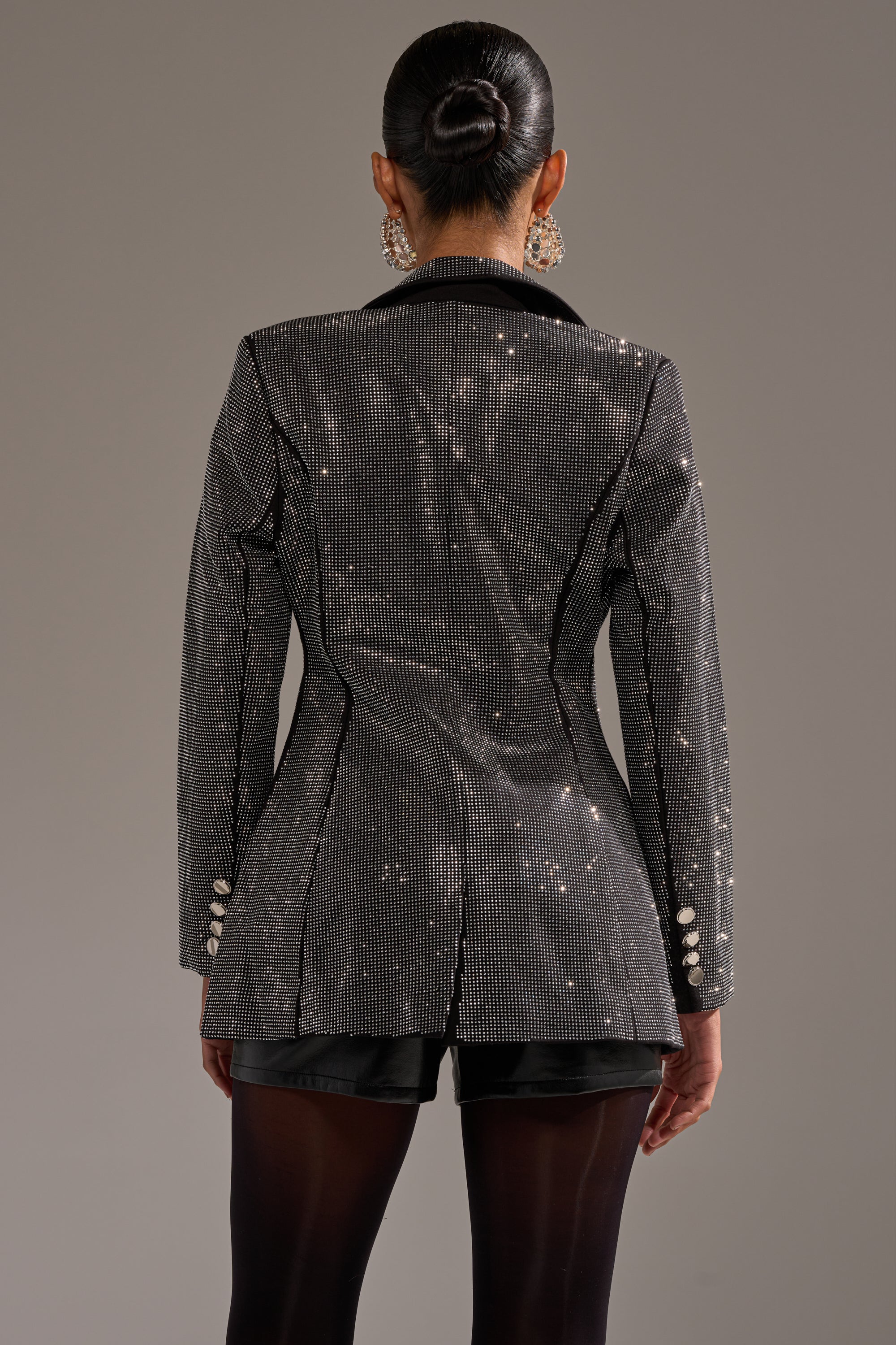 A person with dark hair in a bun models the MORE CHAMPAGNE PLEASE BACKLESS RHINESTONE BLAZER, styled with black shorts, sheer tights, and statement earrings, posing with their back to the camera against a plain background.