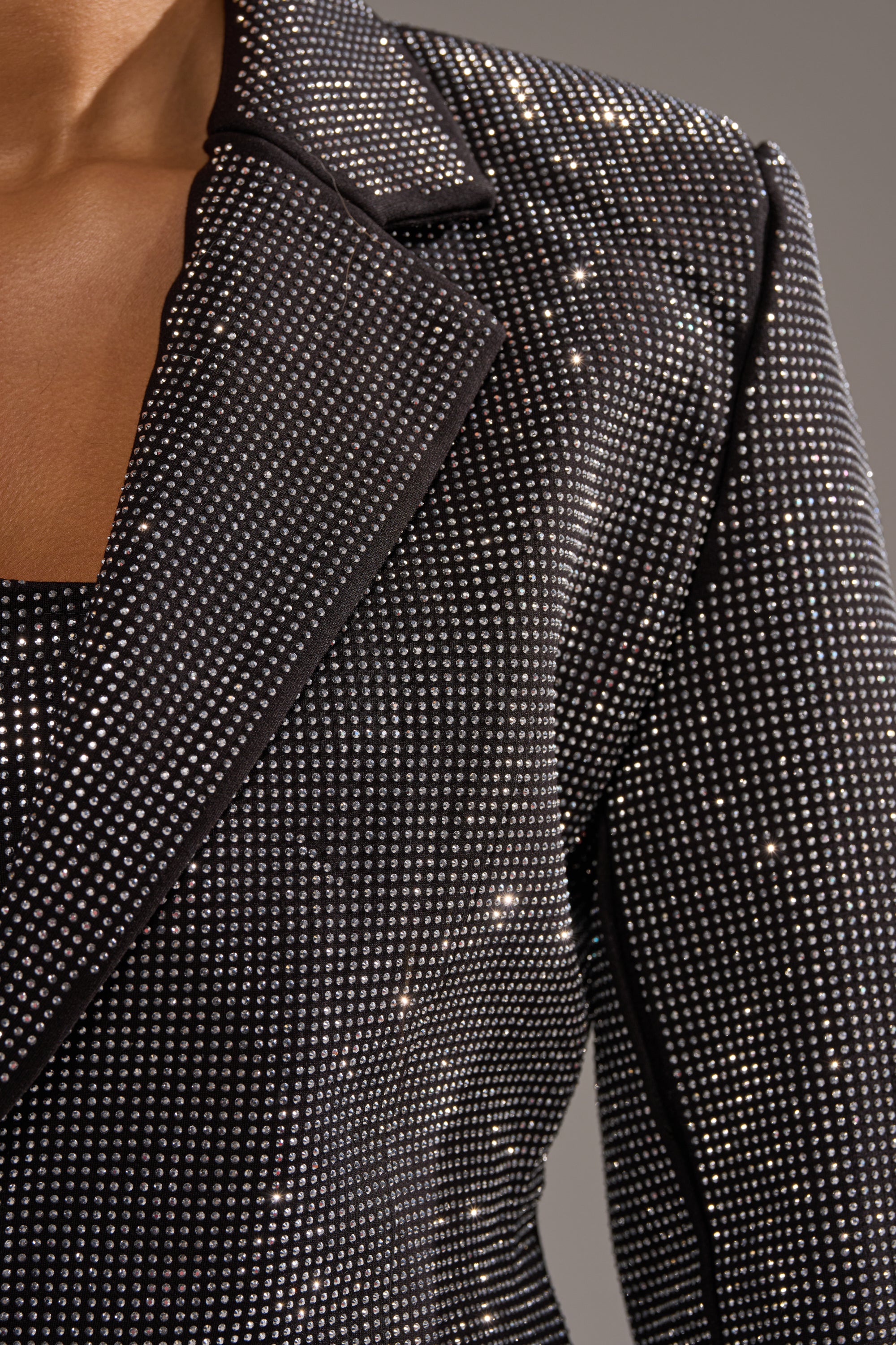 Close-up of a person wearing the MORE CHAMPAGNE PLEASE BACKLESS RHINESTONE BLAZER, a black blazer adorned with small, reflective silver rhinestones in a sparkling dotted pattern against a plain gray background.