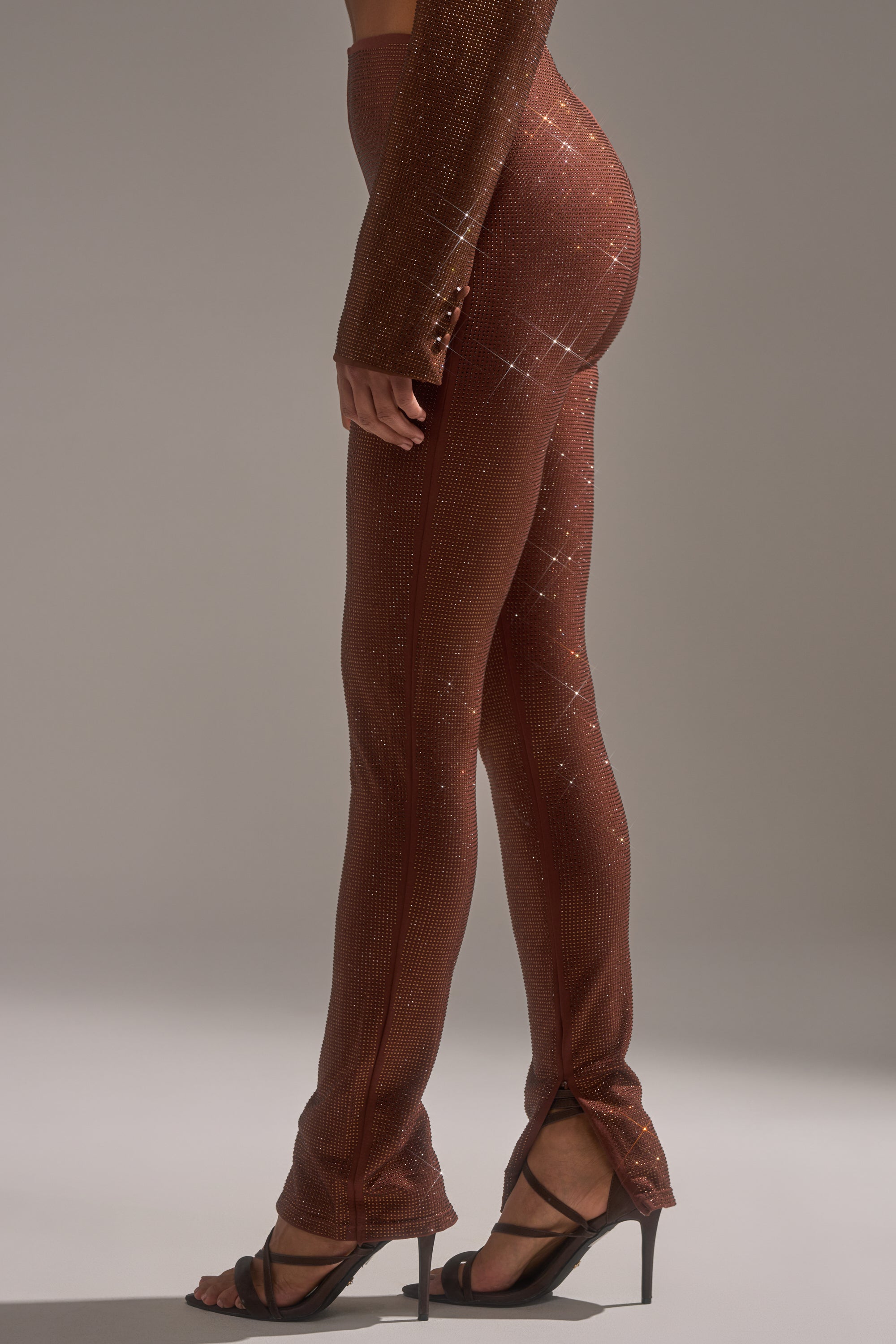 A model displays the MAKE A TOAST RHINESTONE PANT—sparkly, fitted, reddish-brown skinny-leg pants—paired with black high-heeled sandals in a neutral studio, only showing their lower half in profile.