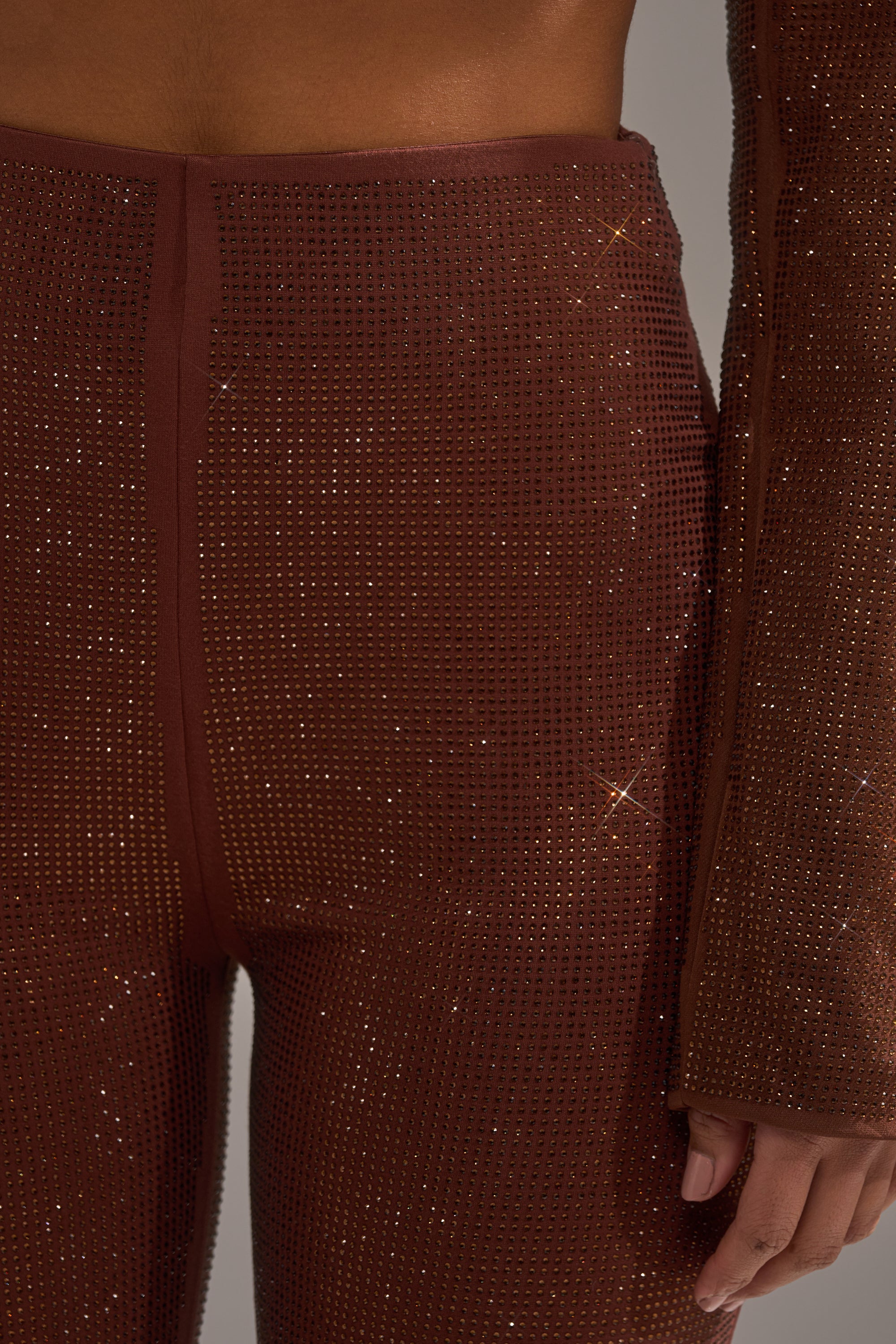 A close-up of a person wearing the MAKE A TOAST RHINESTONE PANT, featuring high-rise, brown, sparkly skinny legs with rhinestone embellishments. Only the lower torso, hip, and hand are visible.