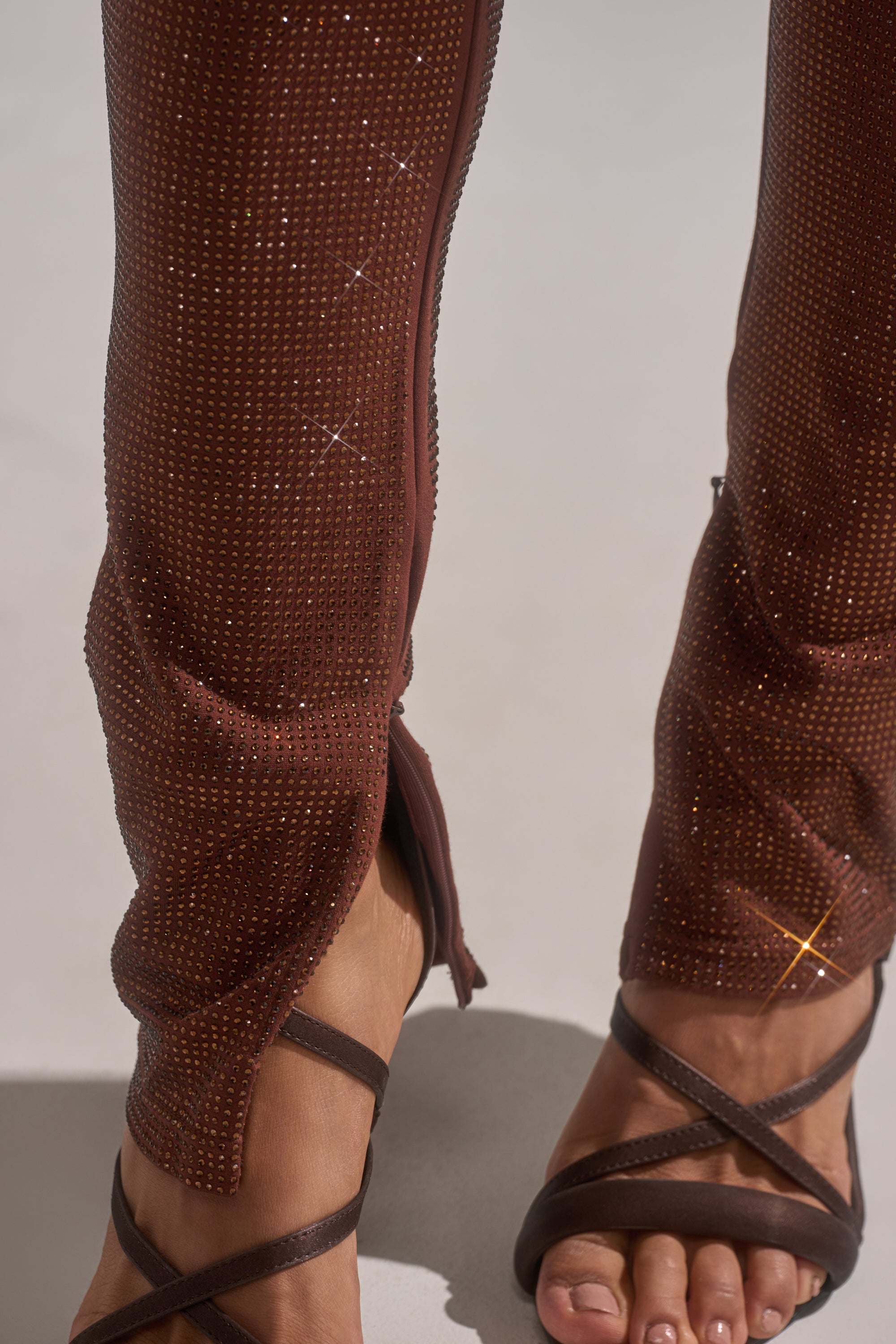 A close-up of lower legs wearing MAKE A TOAST RHINESTONE PANT featuring high-rise skinny fit, sparkling rhinestones, front ankle zippers, and brown strappy sandals. The photo emphasizes the pant’s shimmering texture.