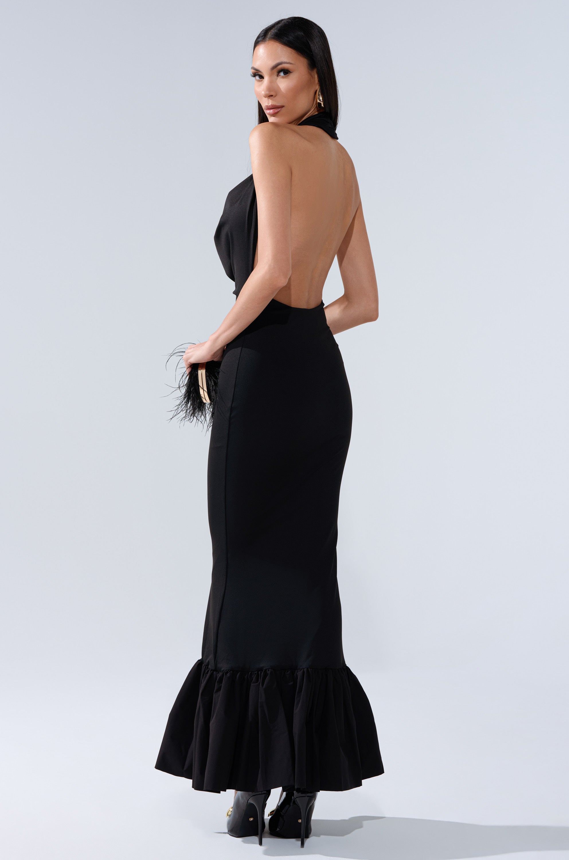 A woman poses in the ELLIE MAXI DRESS, a sleeveless black evening gown with a sheer backless halter neckline and ruffled hem, paired with black high heels against a light gray background.