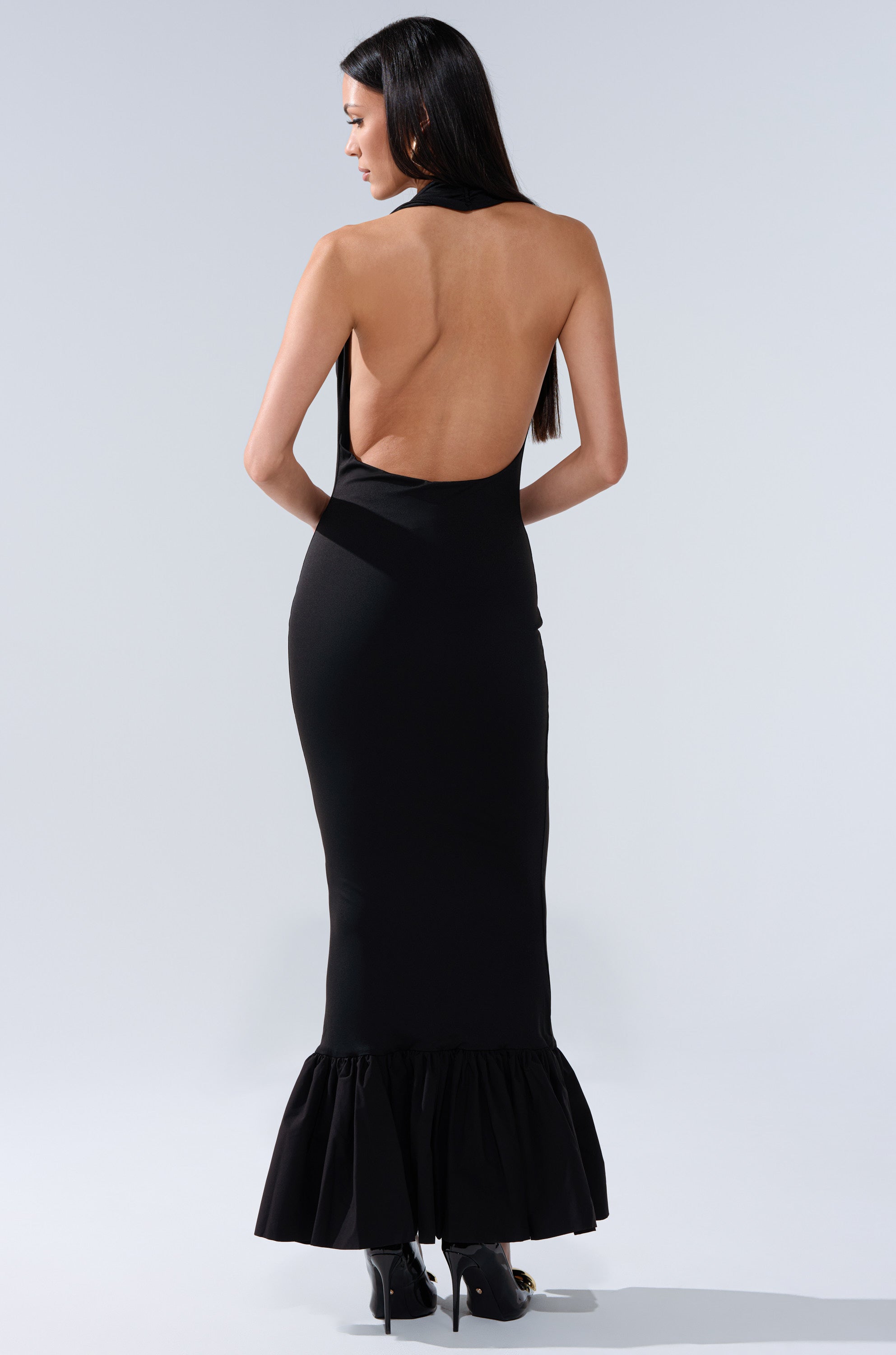 A woman stands facing away in the ELLIE MAXI DRESS, a long backless maxi with a halter neckline and ruffled hem, paired with black heels. Her straight dark hair cascades down her back against a plain light background.