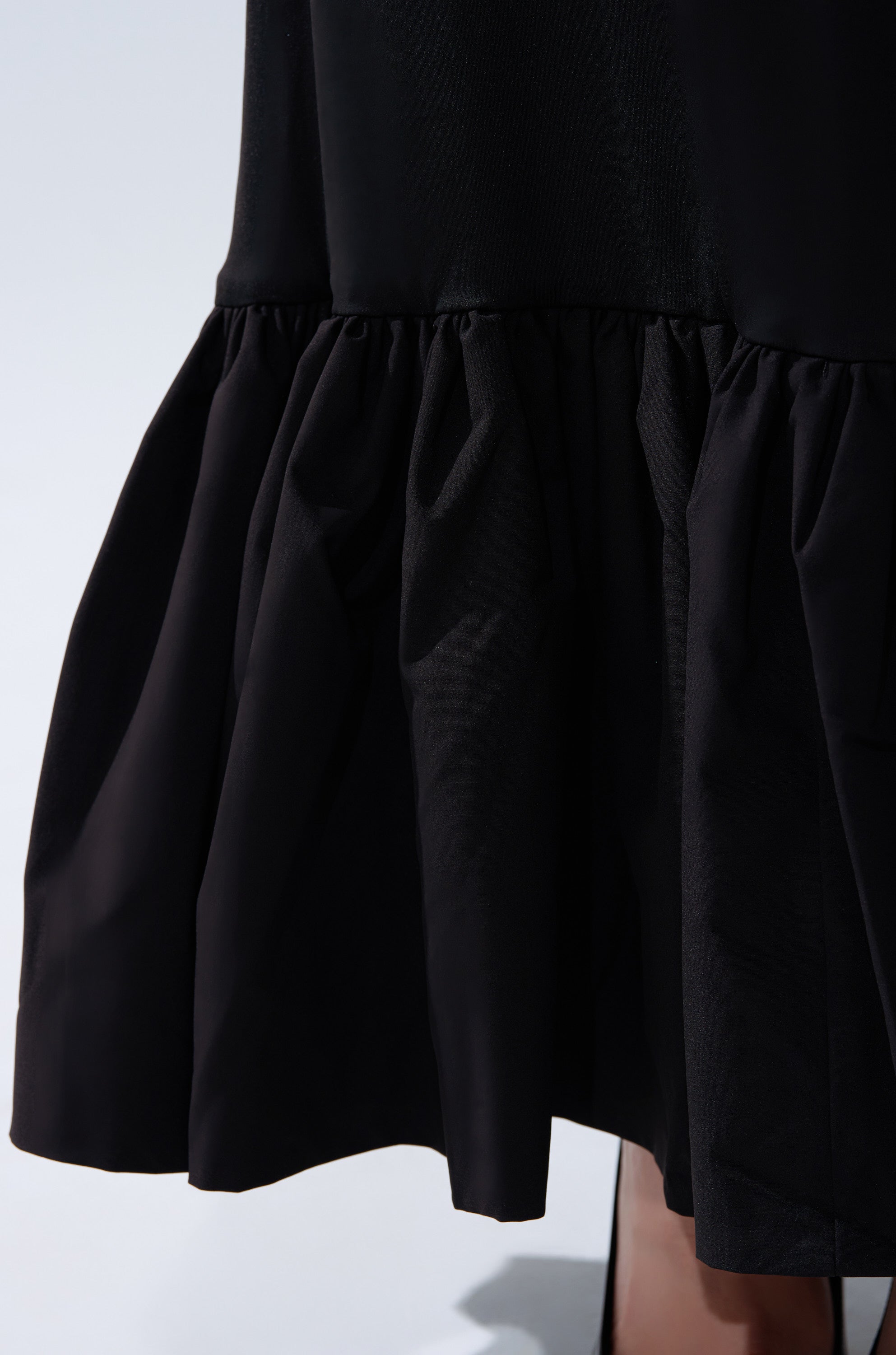 Close-up of the ELLIE MAXI DRESS’s lower part, showcasing its ruffled hem, gentle fabric folds, and subtle sheen against a plain light background.