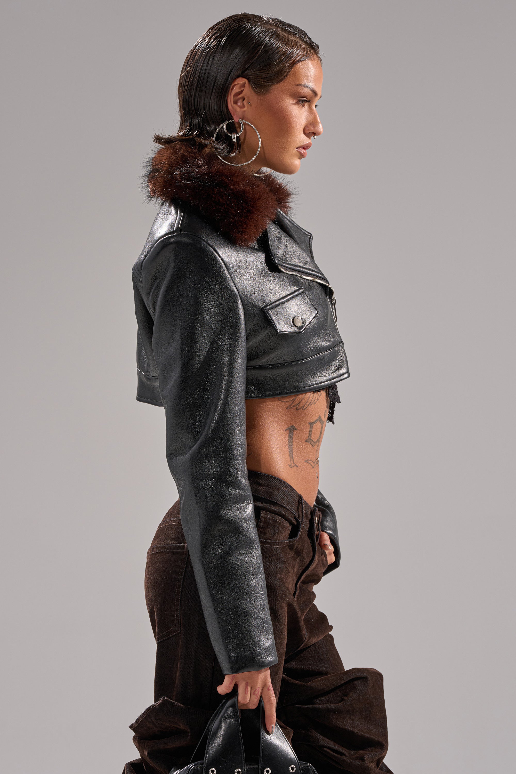 A woman with short, slicked-back hair models the VICTORIA FUR TRIM MOTO, a cropped faux leather jacket with a fur collar, plus hoop earrings and brown pants, revealing a side tattoo against a plain gray background. 4