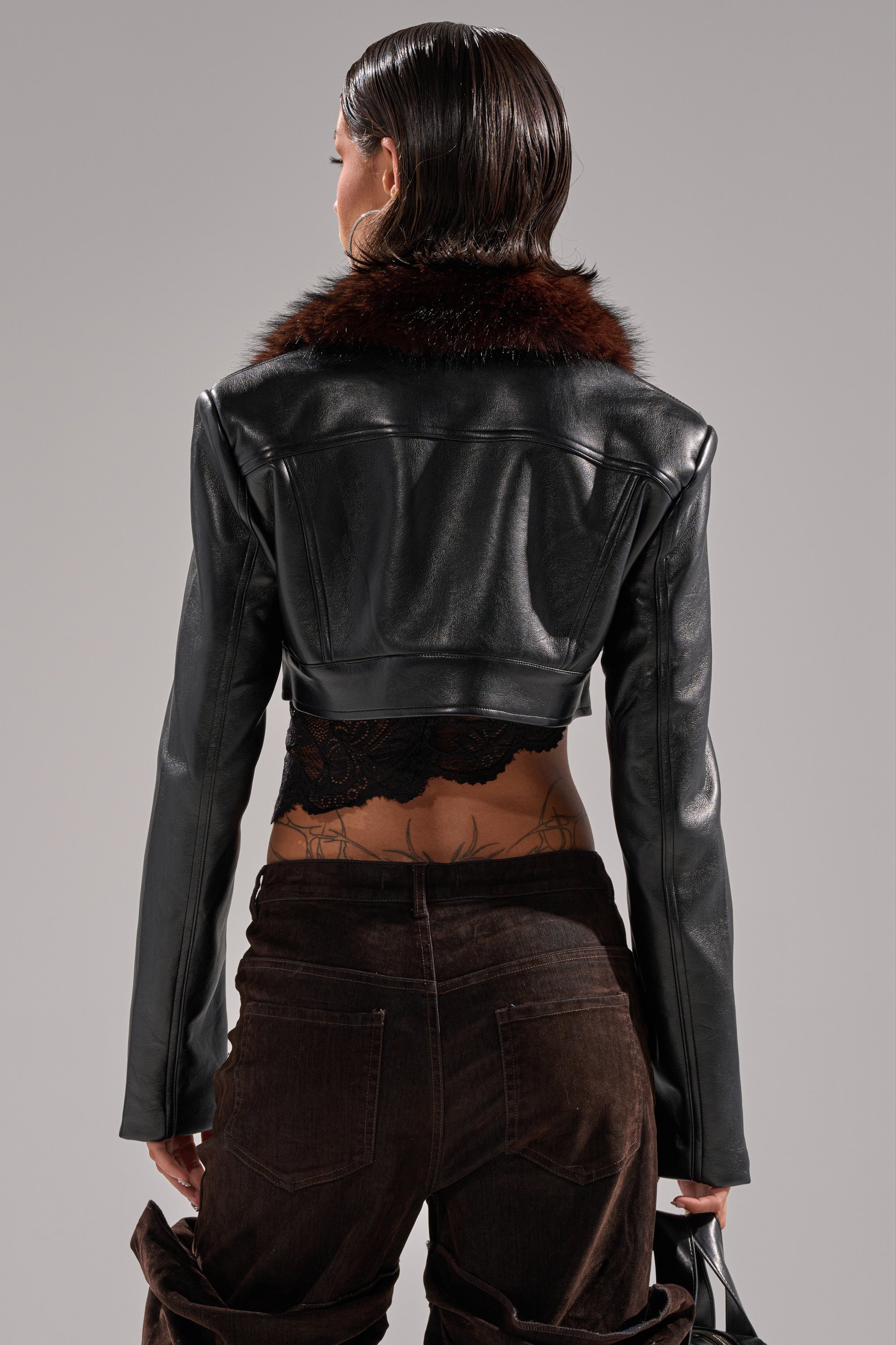 A person with wet, slicked-back hair is seen from behind wearing the VICTORIA FUR TRIM MOTO jacket, featuring a faux leather finish and brown fur collar, styled with a lace top and brown velvety pants revealing lower back tattoos. 5