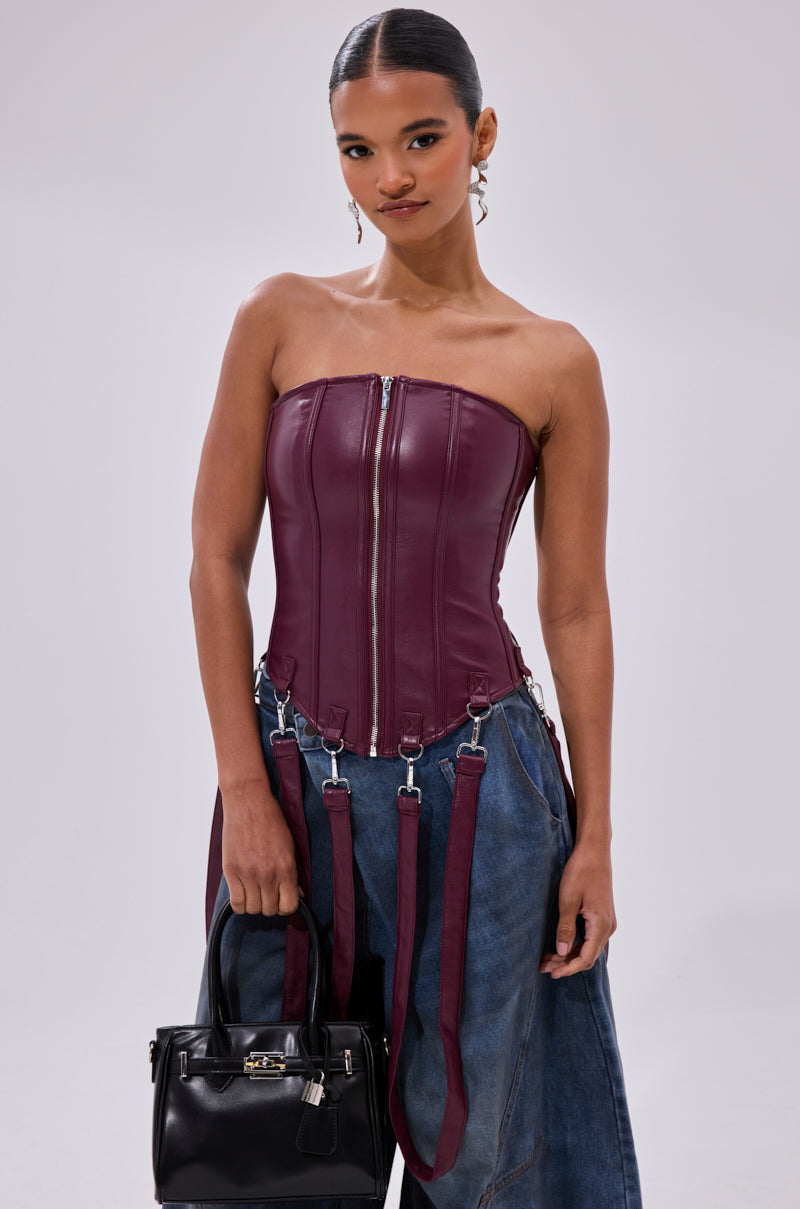 A woman wearing the FAN FAVORITE FAUX LEATHER CORSET with zipper and suspender details, paired with wide-leg blue jeans and holding a black handbag, stands against a plain light background.