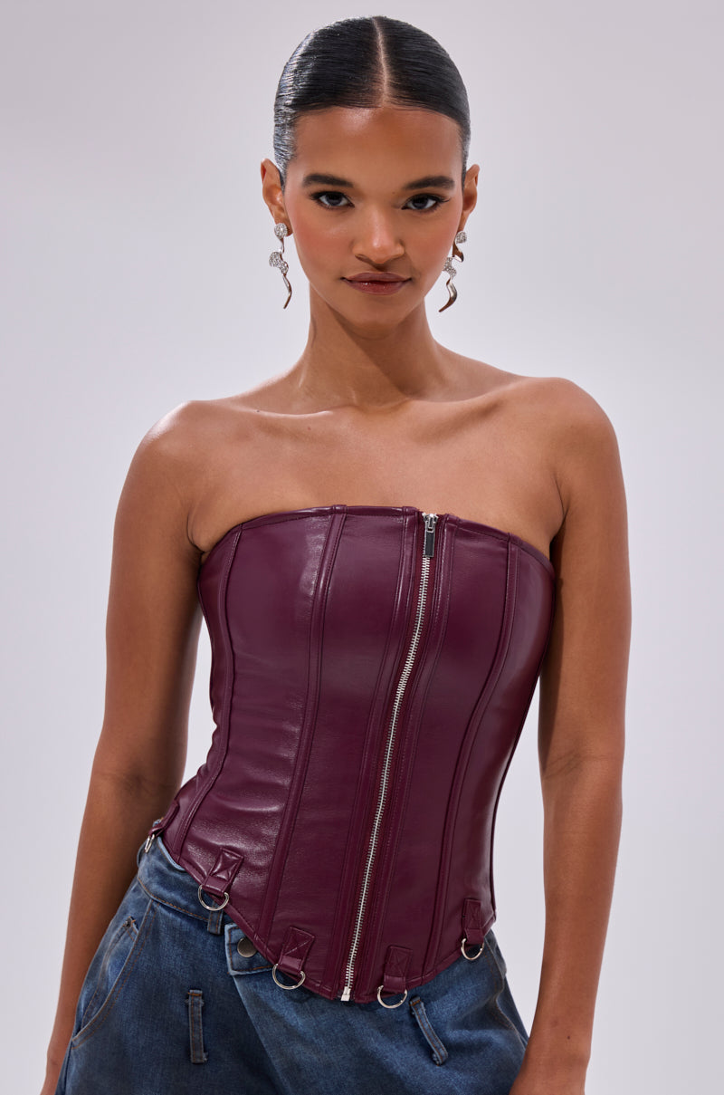 A woman with slicked-back hair confidently models the FAN FAVORITE FAUX LEATHER CORSET, a strapless zipper top, paired with blue jeans and statement earrings against a plain light background.