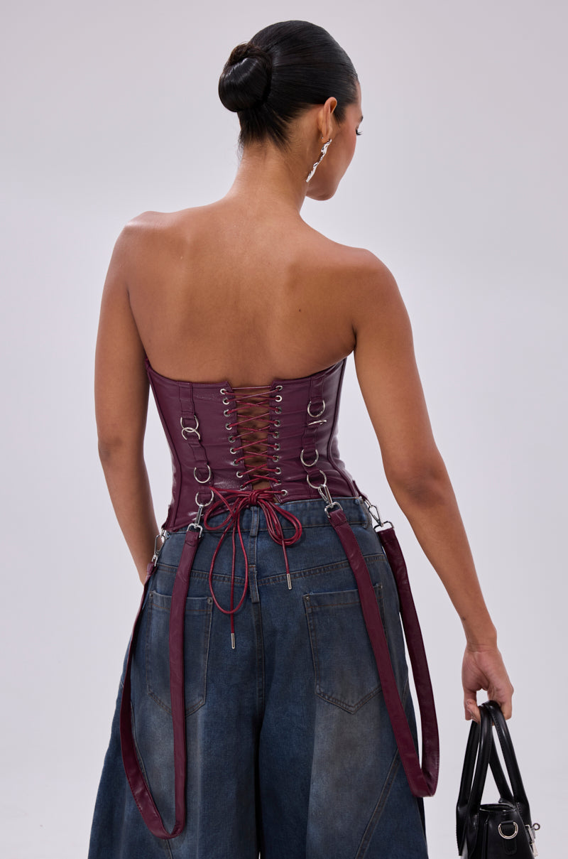 A woman with dark hair in a bun wears the FAN FAVORITE FAUX LEATHER CORSET featuring metal ring details and a lace-up back, paired with high-waisted wide-leg blue jeans. Holding a black handbag, she faces away against a plain background.