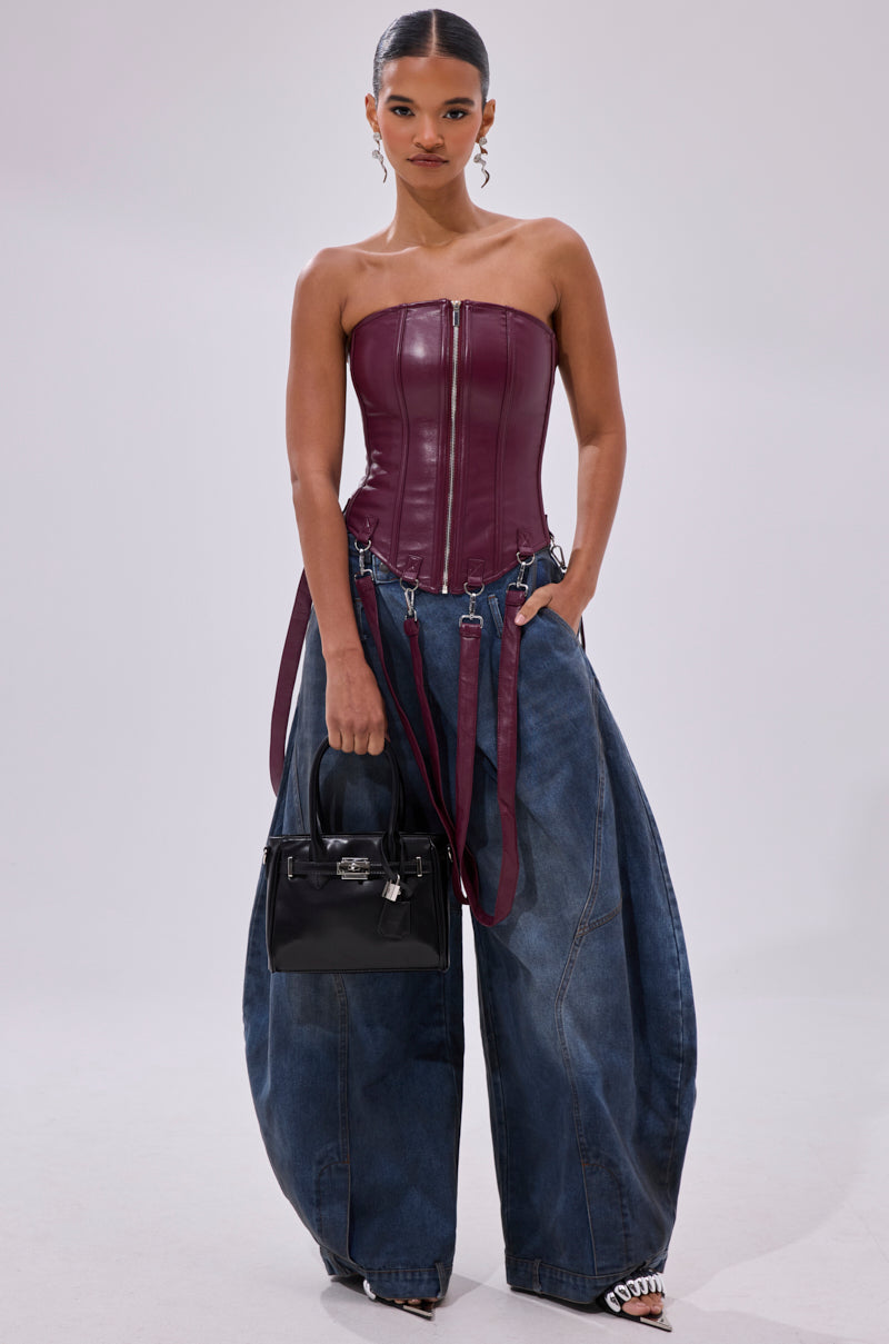 A woman stands confidently in the FAN FAVORITE FAUX LEATHER CORSET with a front zipper, styled with oversized blue denim jeans, black heels, a small black handbag, and her hair in a sleek ponytail.