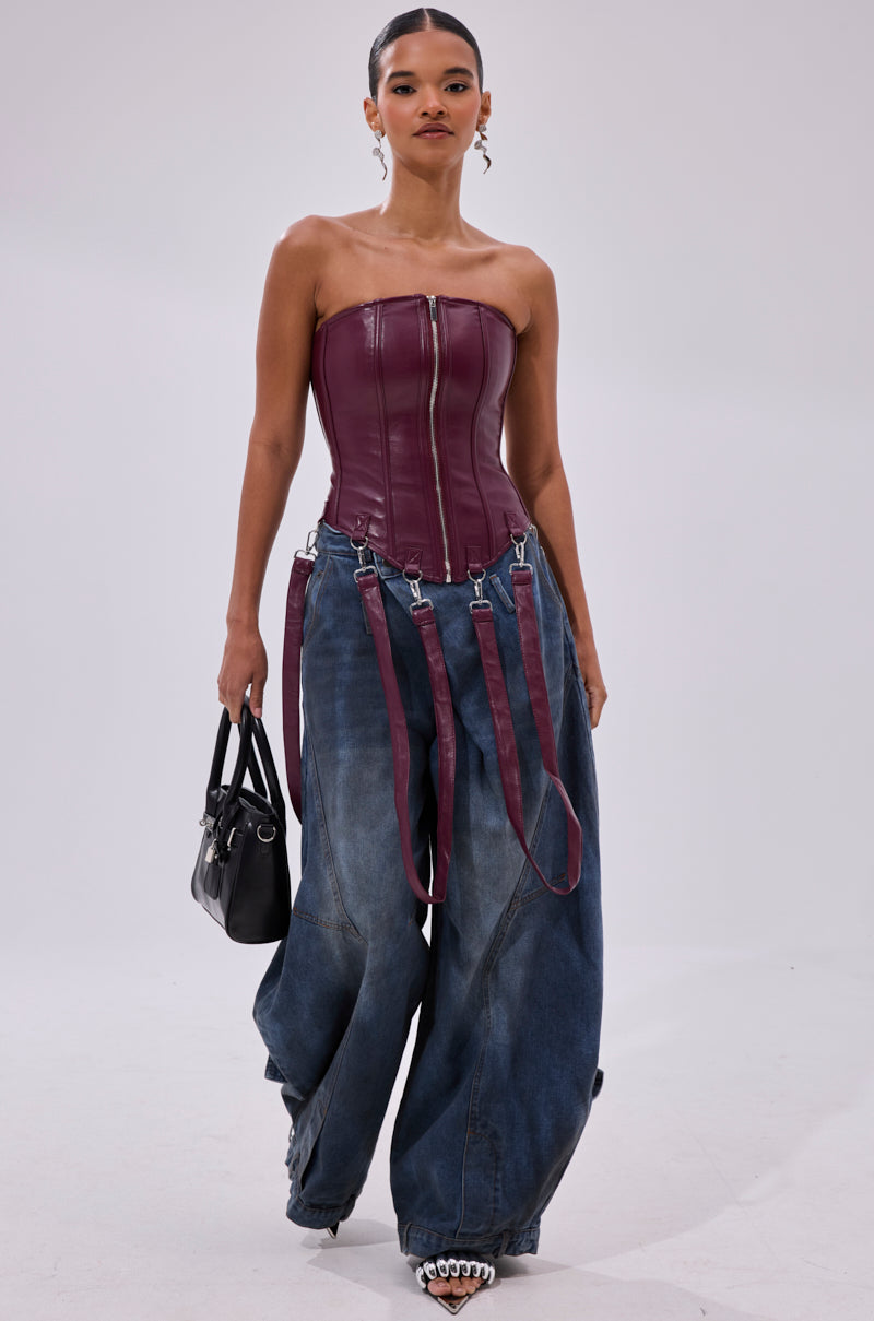 A woman models the FAN FAVORITE FAUX LEATHER CORSET with a metallic zipper, paired with oversized blue jeans featuring burgundy straps, a black handbag, and pointed shoes against a plain light background.