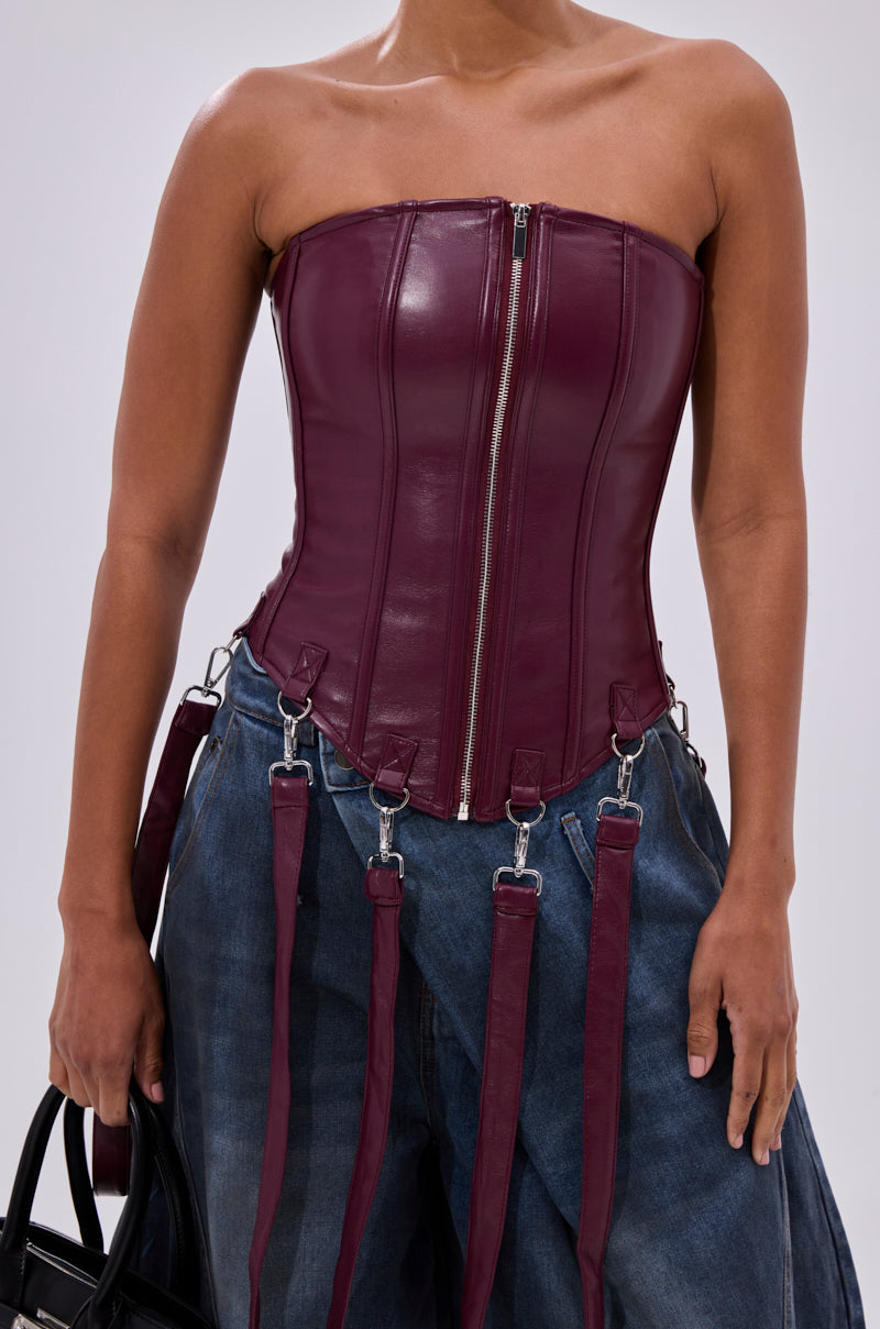 Wearing the FAN FAVORITE FAUX LEATHER CORSET—a strapless faux leather top with a front zipper and detachable harness straps—paired with loose blue jeans and holding a black handbag.
