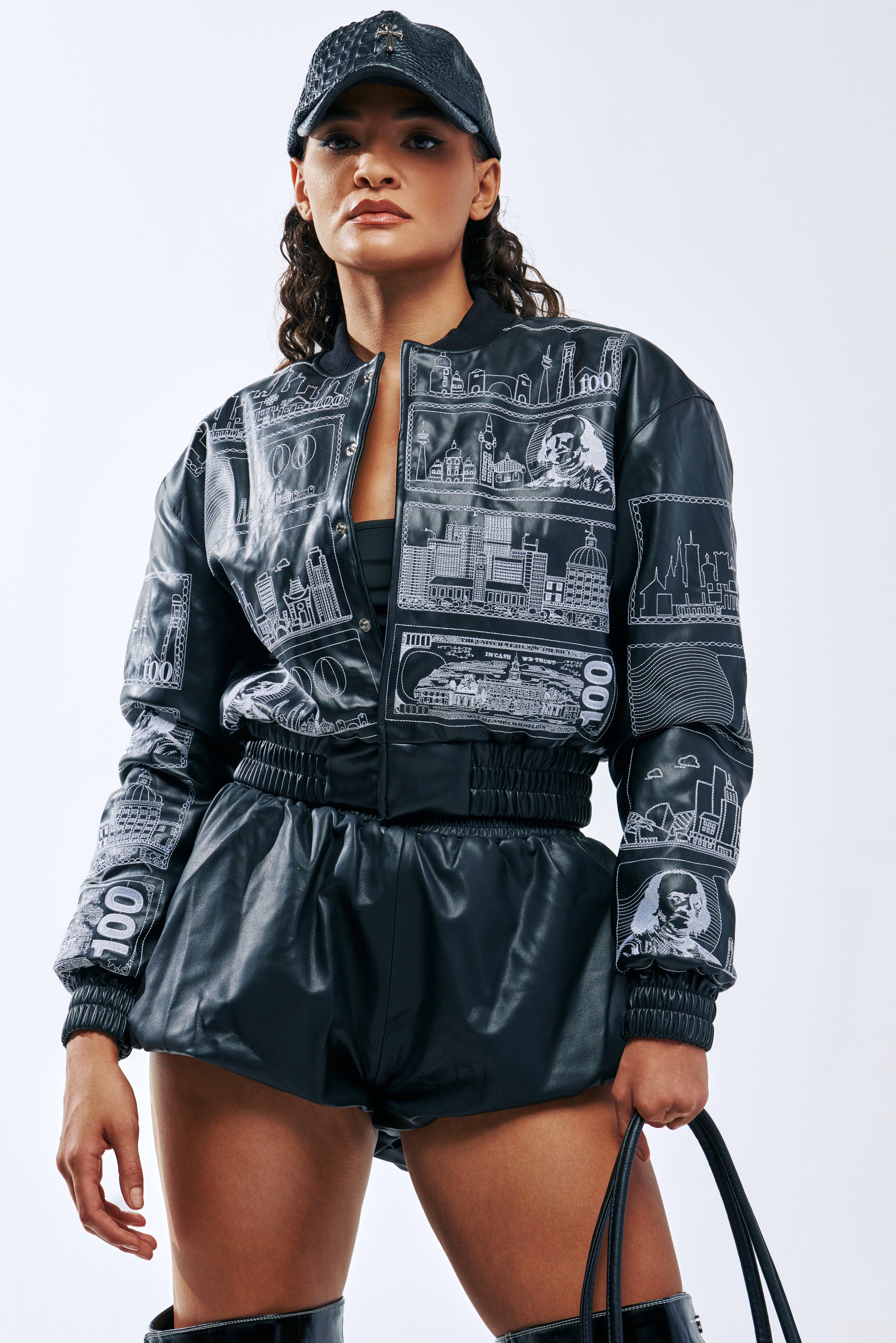 A model wearing the MONEY IS THE MOTIVE EMBROIDERED BOMBER in black, featuring white embroidered city skylines and currency-inspired graphics, paired with a matching black baseball cap and shorts.