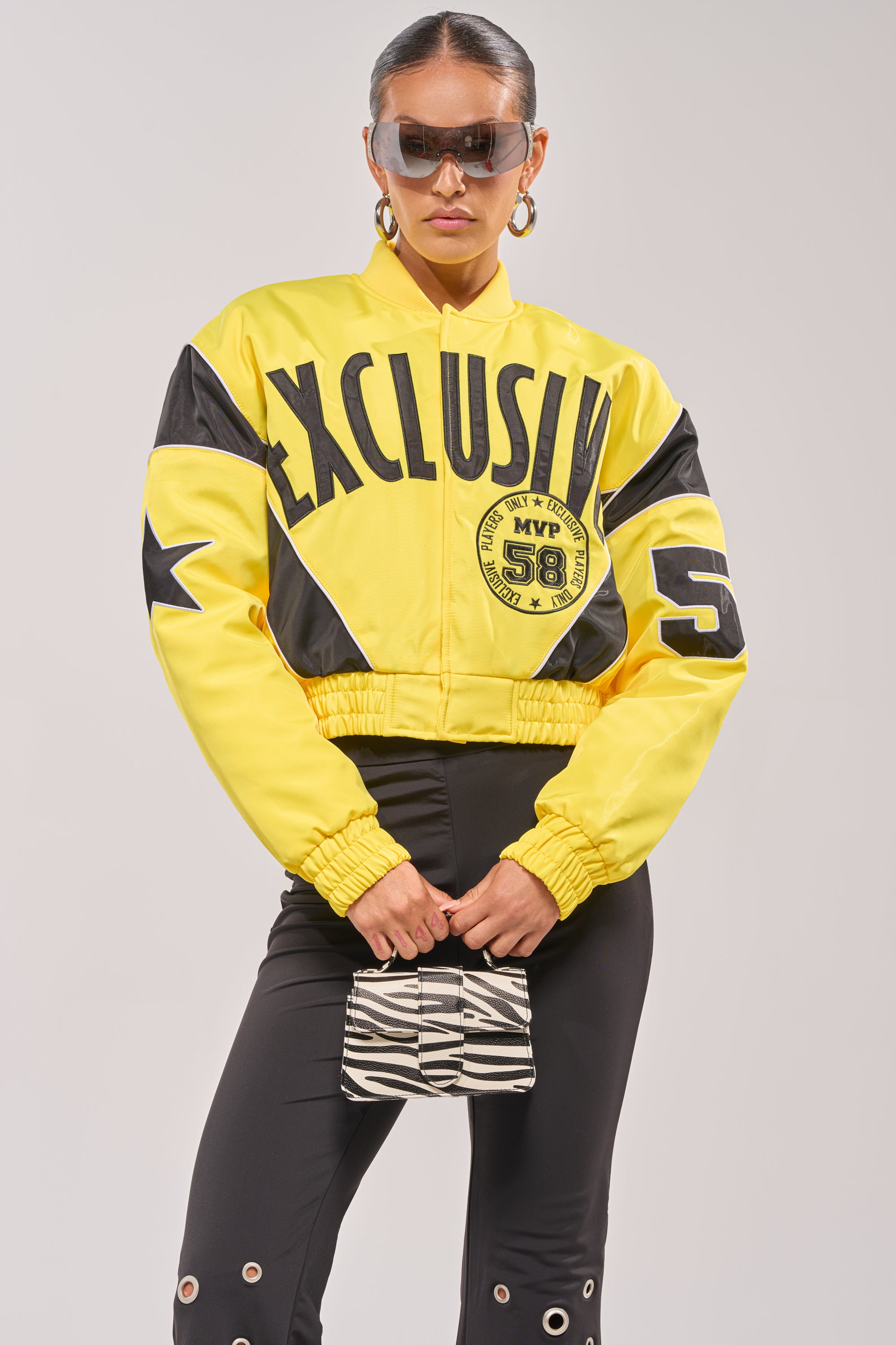 A woman rocks the BILLIE EMBROIDERED NYLON BOMBER, bold and yellow with "EXCLUSIVE" and "MVP 58," styled with grommet-detailed black pants, large sunglasses, hoop earrings, and a zebra-print bag—ultimate street style. 0