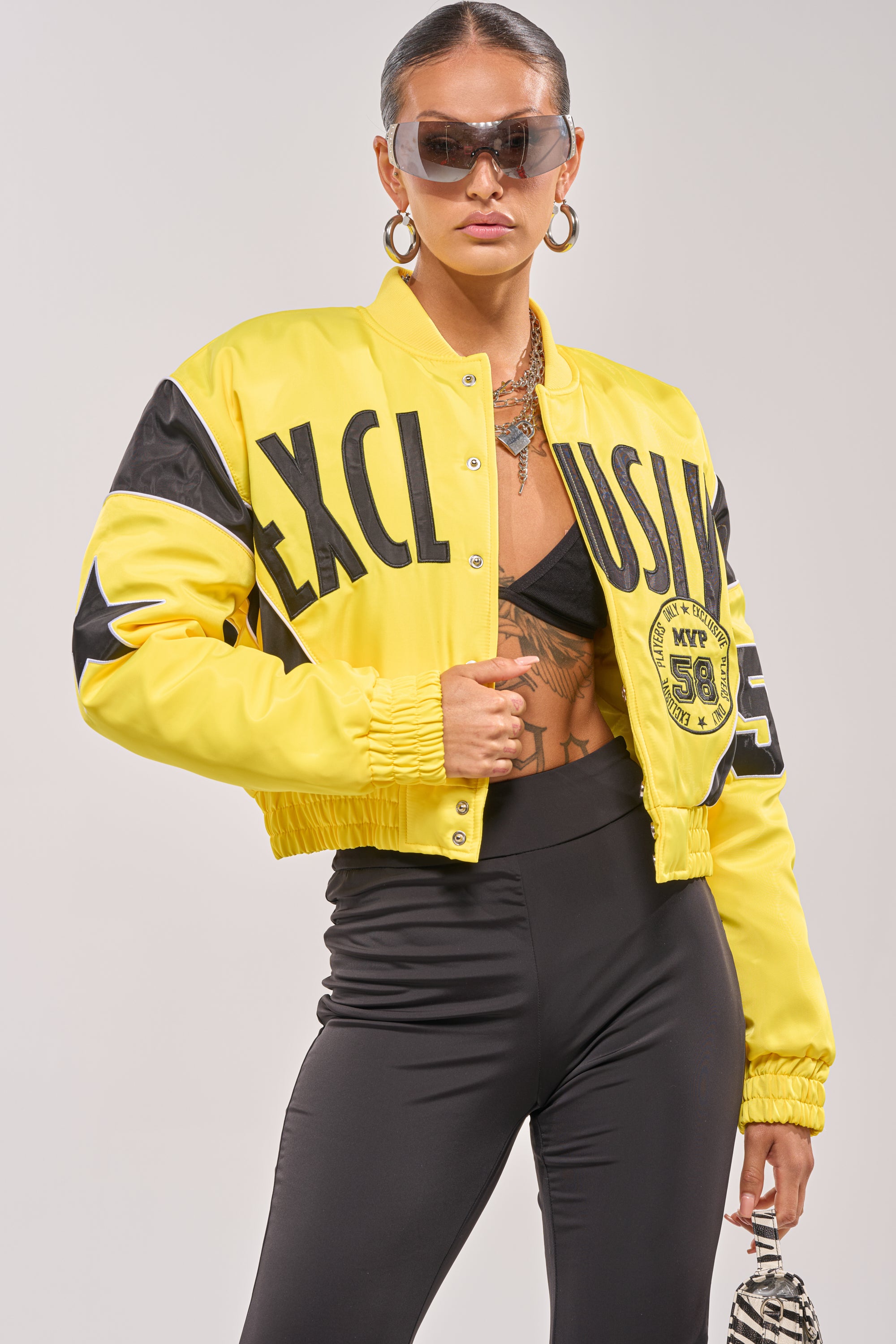 A woman confidently poses in the BILLIE EMBROIDERED NYLON BOMBER, styled with large sunglasses, a black top, black leggings, and a small patterned bag. 1