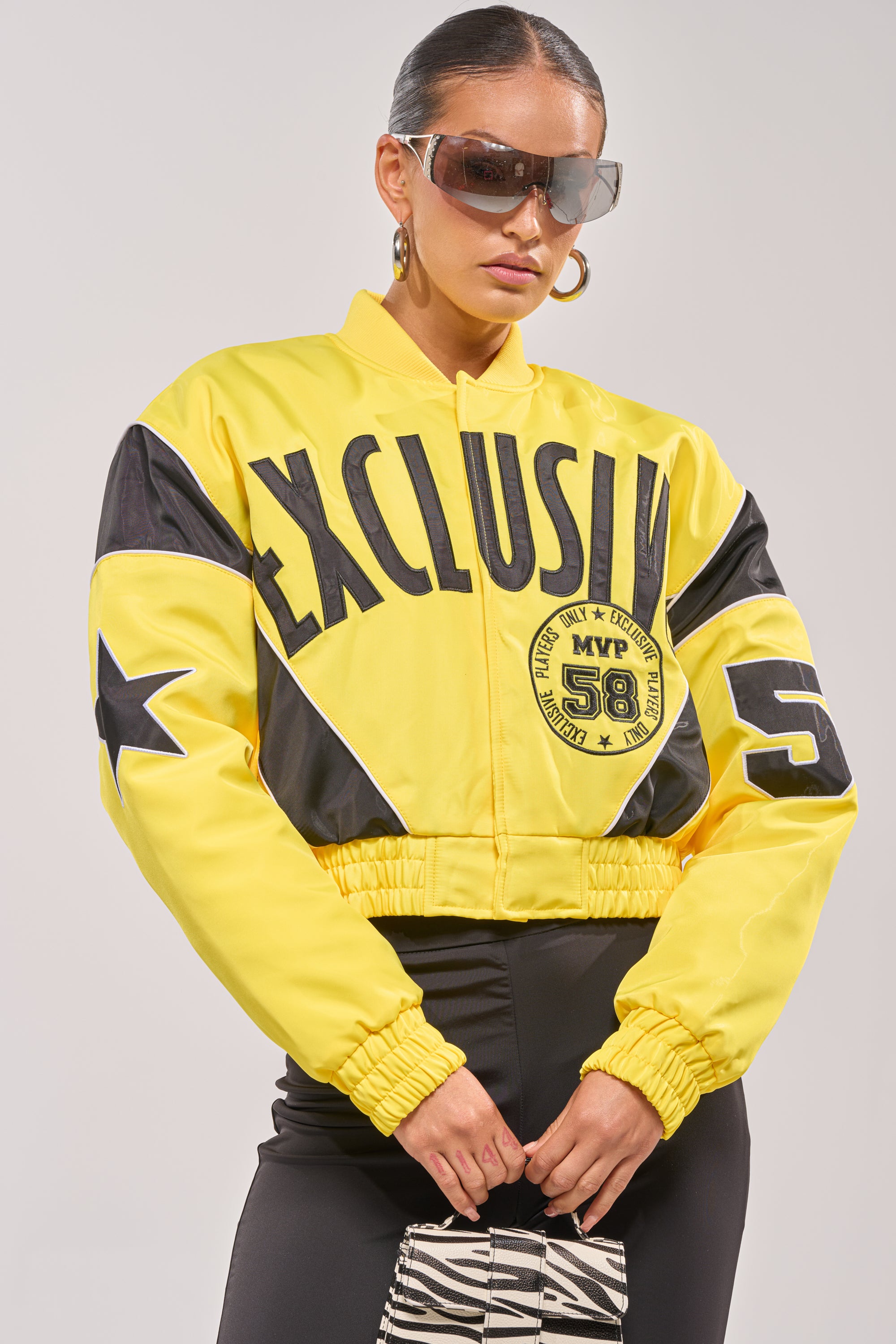 A woman in the BILLIE EMBROIDERED NYLON BOMBER—a yellow varsity jacket with bold "EXCLUSIVE," stars, and "58"—paired with black pants, large sunglasses, and a black-and-white patterned purse. 2