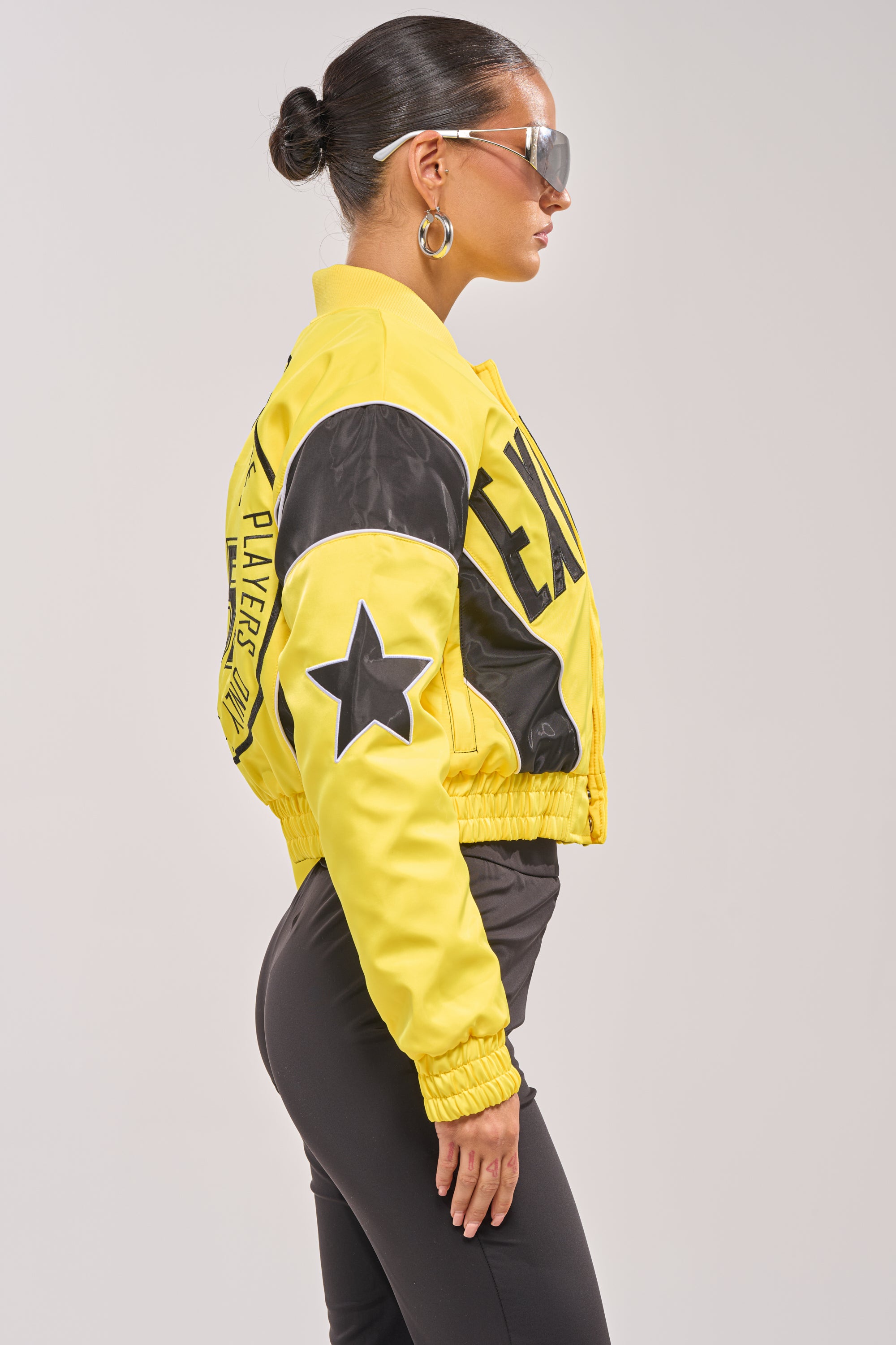 A woman in profile wears the BILLIE EMBROIDERED NYLON BOMBER— a yellow cropped jacket with bold lettering and a star patch on the sleeve—paired with black pants, hoop earrings, and sunglasses against a plain background. 3
