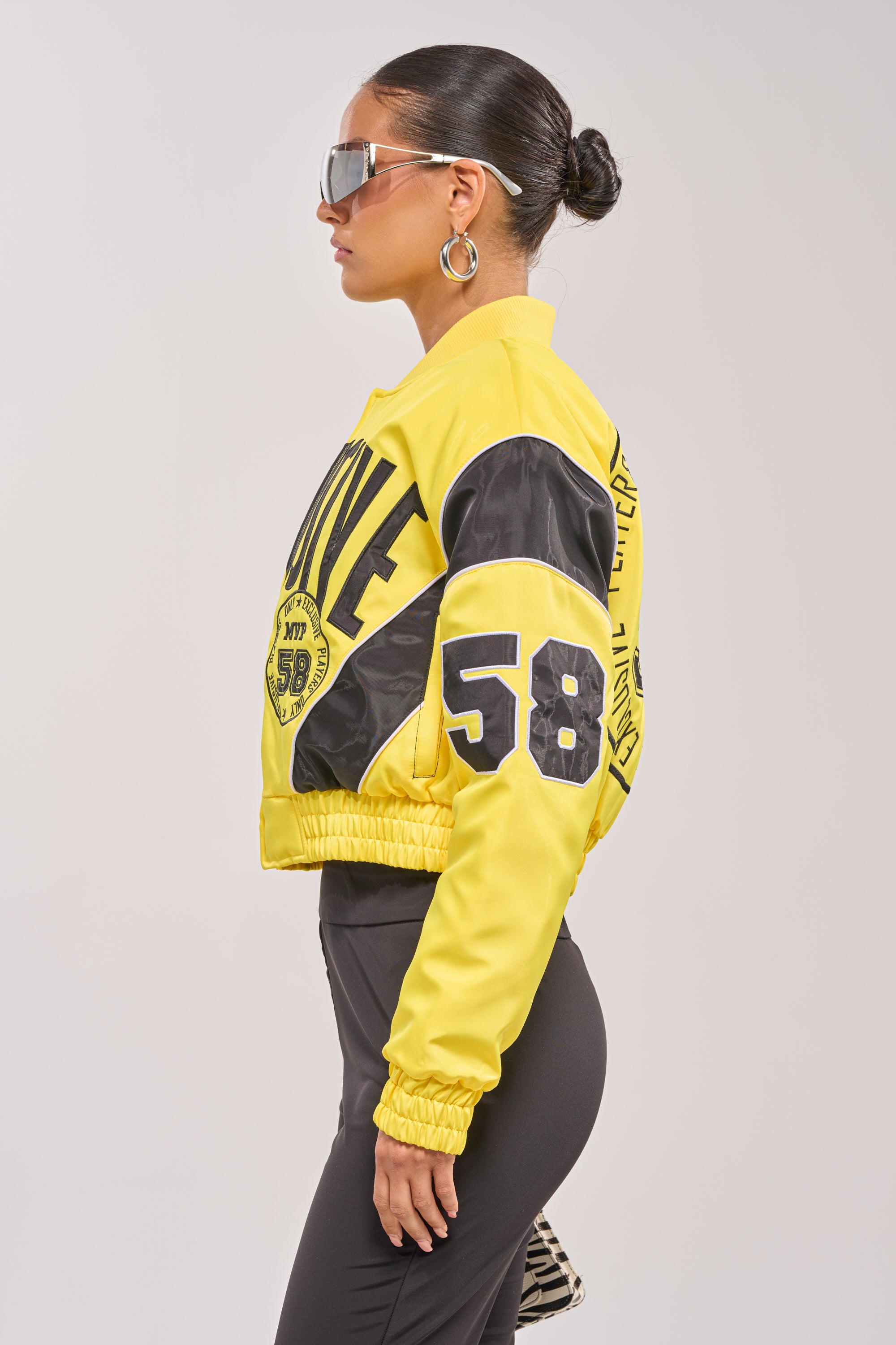 A woman in profile wears large sunglasses, hoop earrings, and the BILLIE EMBROIDERED NYLON BOMBER—a yellow varsity jacket with black details, "58" on the sleeve, bold text on the back. Her dark hair is styled in a sleek bun. 4