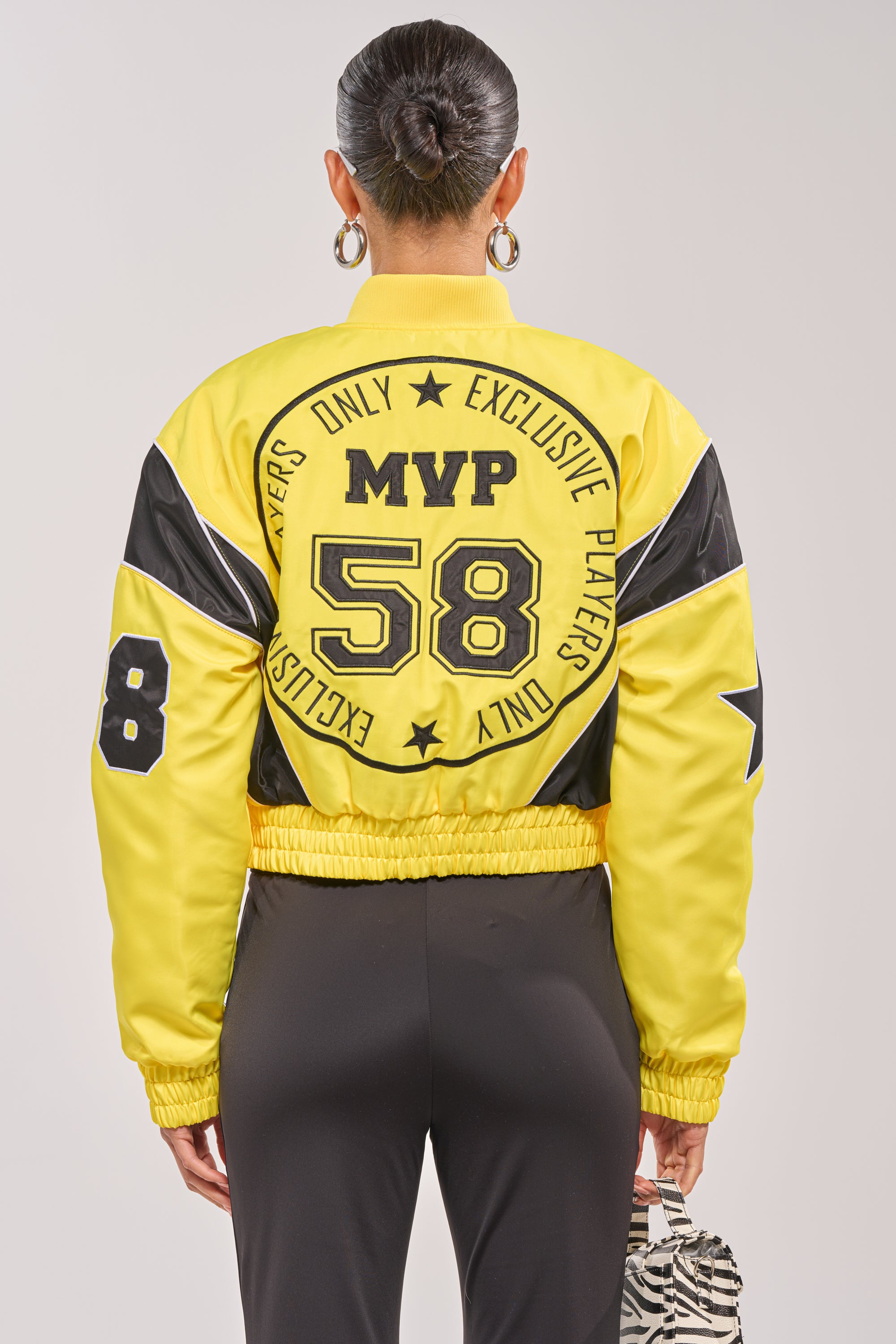 A person with hair in a bun wears the BILLIE EMBROIDERED NYLON BOMBER, a yellow varsity jacket with "MVP 58" and "Exclusive Players Only" on the back. They carry a zebra-print bag and wear large hoop earrings. 5