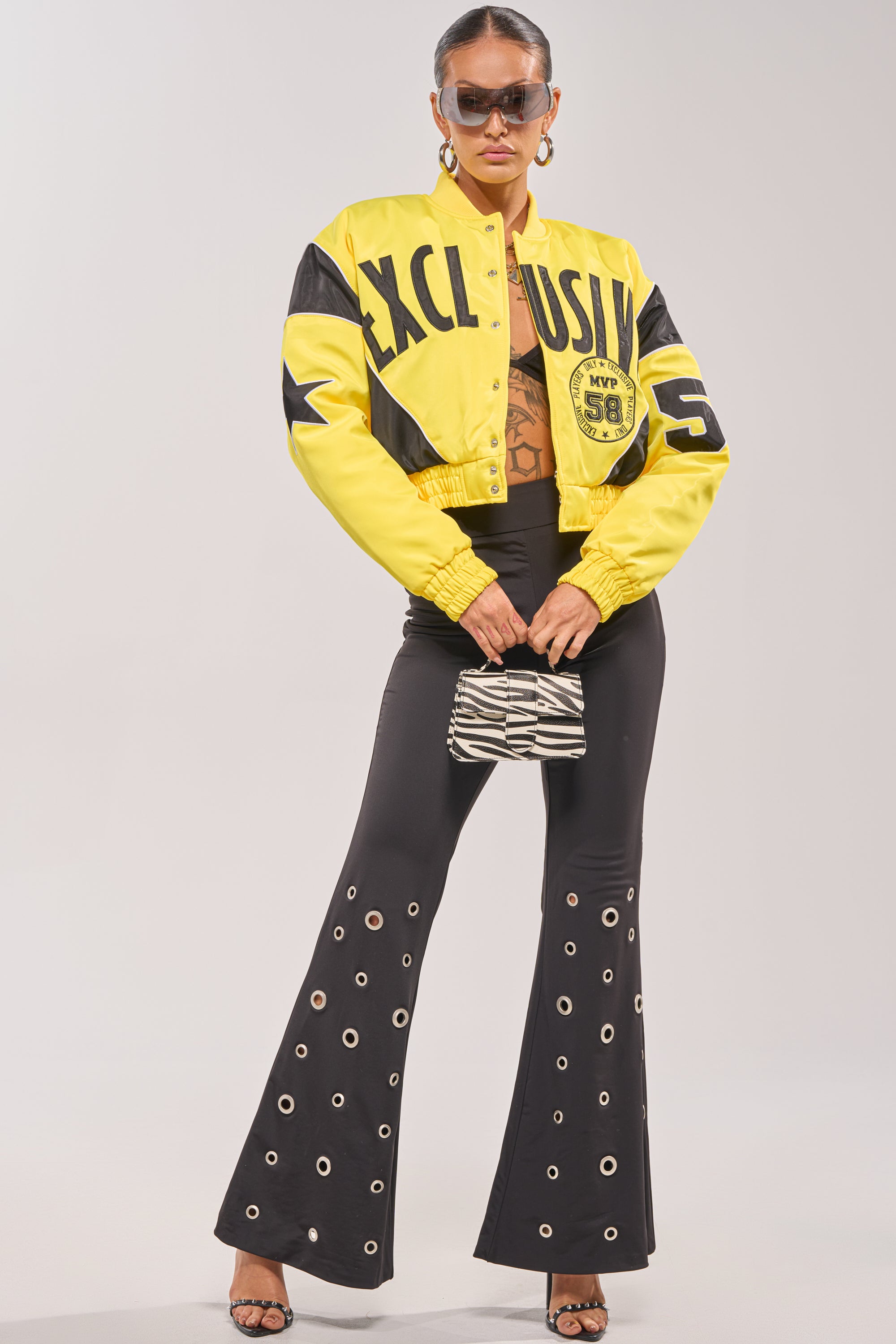 A person wears the BILLIE EMBROIDERED NYLON BOMBER in bold yellow with "EXCLUSIVE" lettering, paired with black flared grommet pants, black heels, sunglasses, hoop earrings, and a small zebra-print handbag. 6
