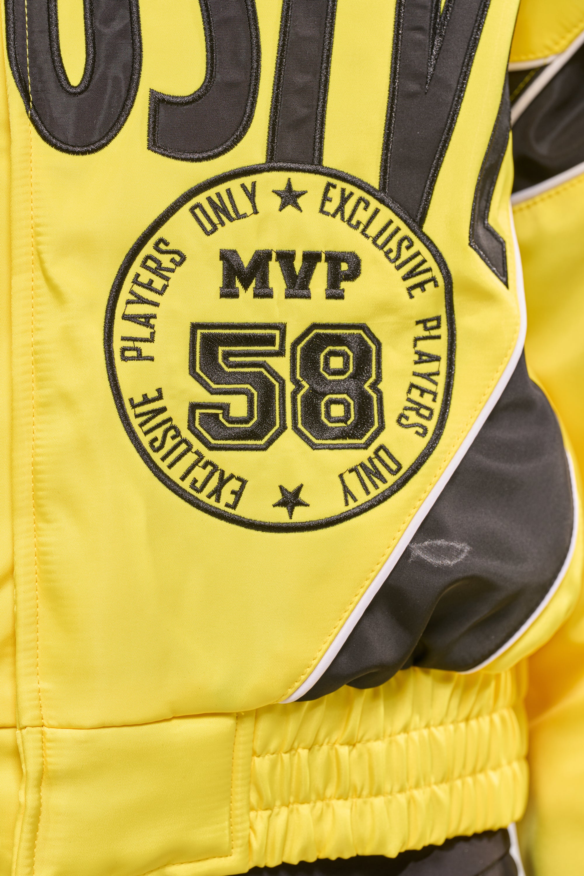 A close-up of the BILLIE EMBROIDERED NYLON BOMBER, a yellow nylon jacket featuring a circular patch with "MVP 58" and "Exclusive Players Only" embroidered in black. 7