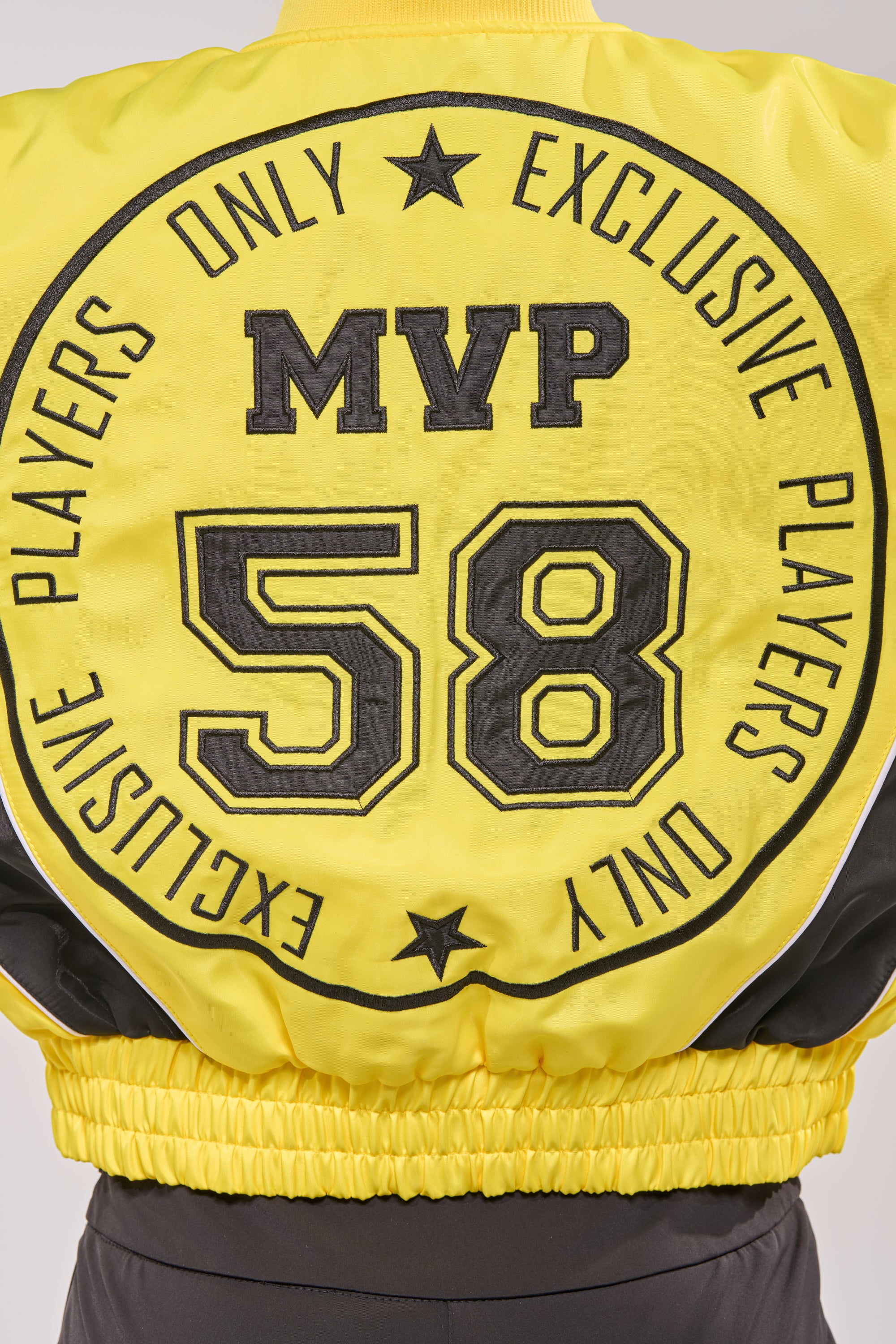 Back of the BILLIE EMBROIDERED NYLON BOMBER in bright yellow with bold black "EXCLUSIVE PLAYERS ONLY," "MVP," and "58" text, star and circular details, plus an elastic waistband. 8