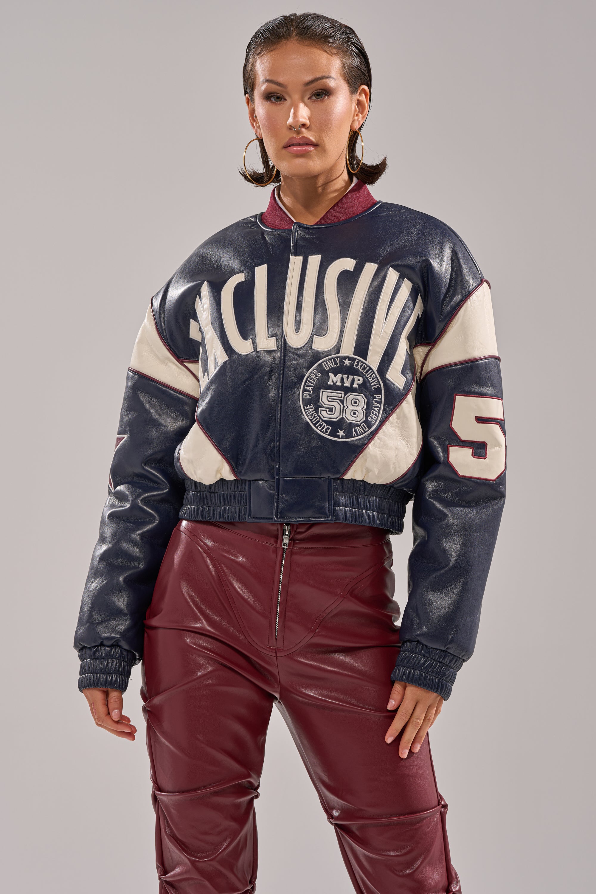 A woman stands confidently against a gray background, wearing large hoop earrings, high-waisted maroon leather pants, and the BUTTER LUXE VARSITY BOMBER—navy blue and white with "EXCLUSIVE" and "58" text on the front.