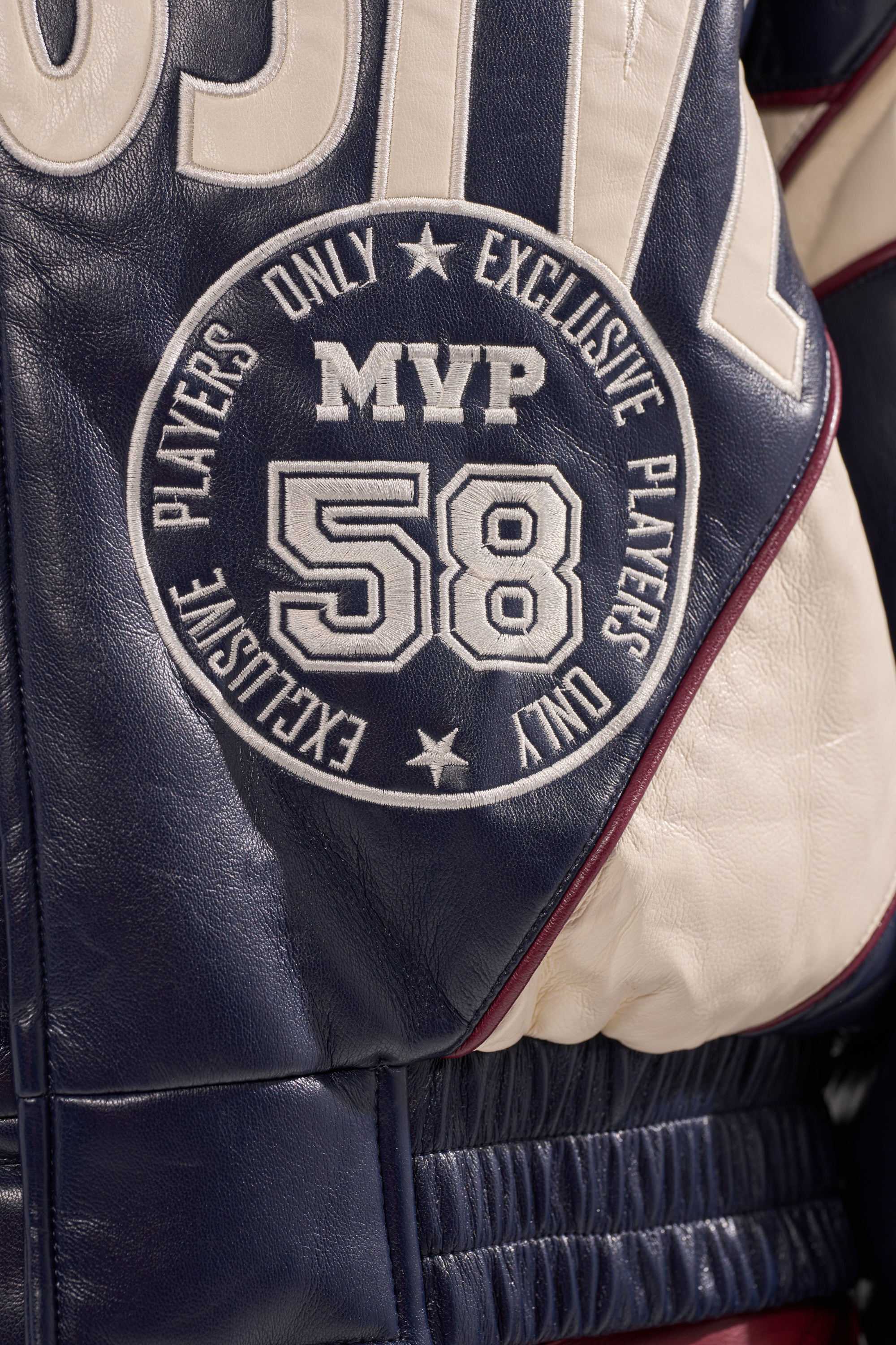 Close-up of a navy blue and white BUTTER LUXE VARSITY BOMBER featuring a patch with "MVP 58" and "Exclusive Players Only" in a circular design around the numbers.