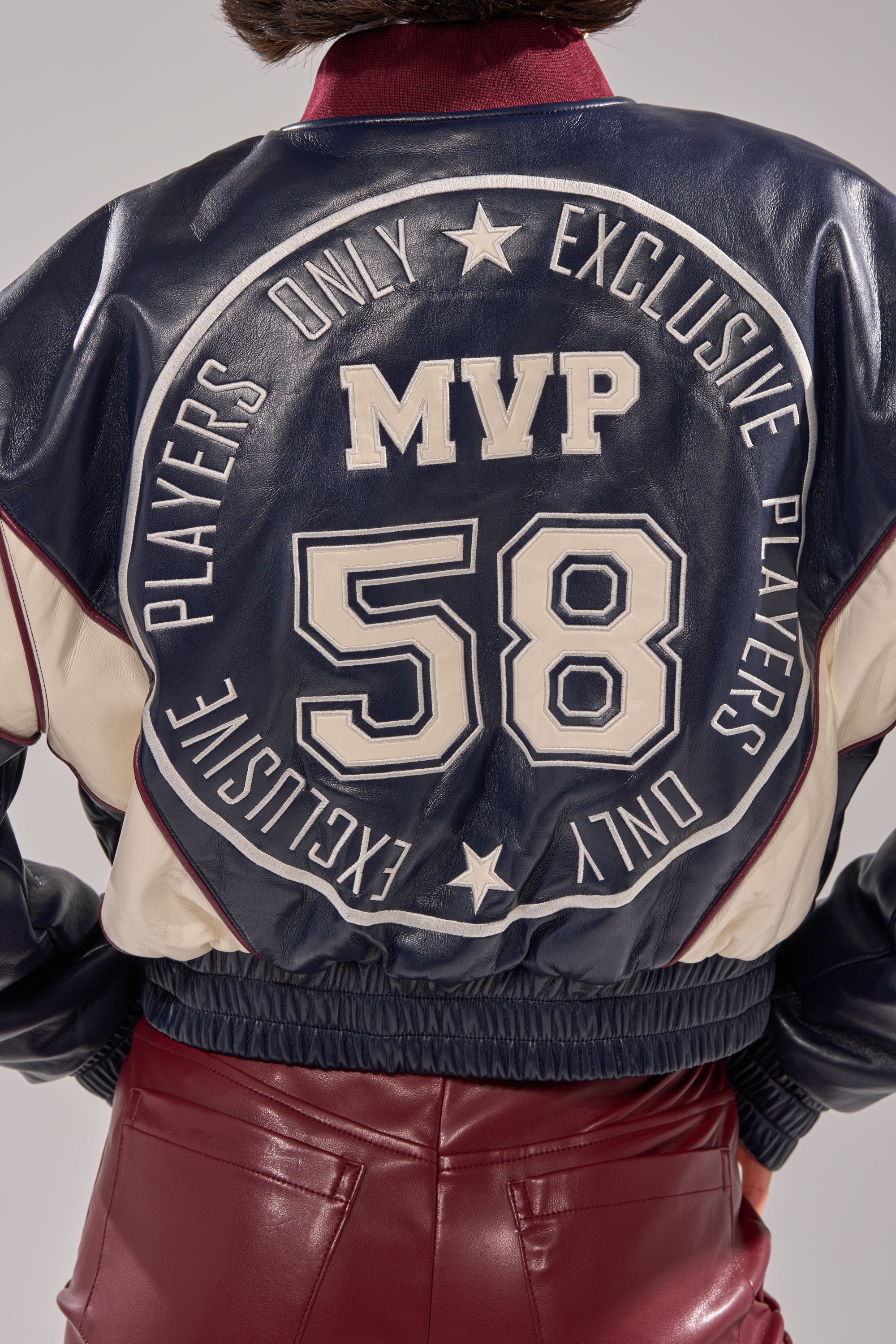 Wearing the BUTTER LUXE VARSITY BOMBER with "MVP 58" and "EXCLUSIVE PLAYERS ONLY" on the back, paired with maroon leather pants for a bold, standout look.