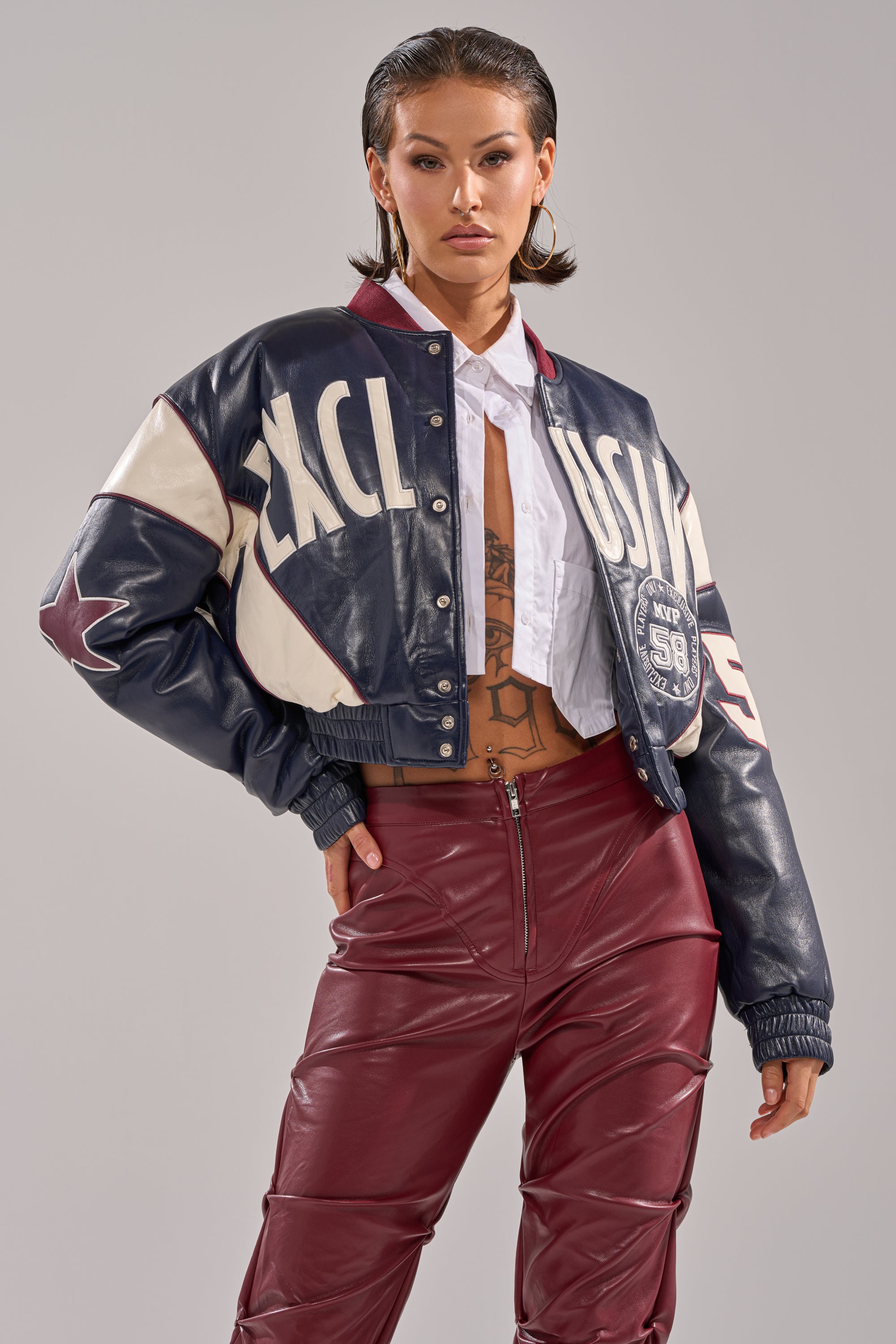 A woman poses confidently in the BUTTER LUXE VARSITY BOMBER, styled over a white cropped shirt with high-waisted burgundy leather pants and gold hoops. Her slicked-back hair and chest tattoo complete the look against a plain background.