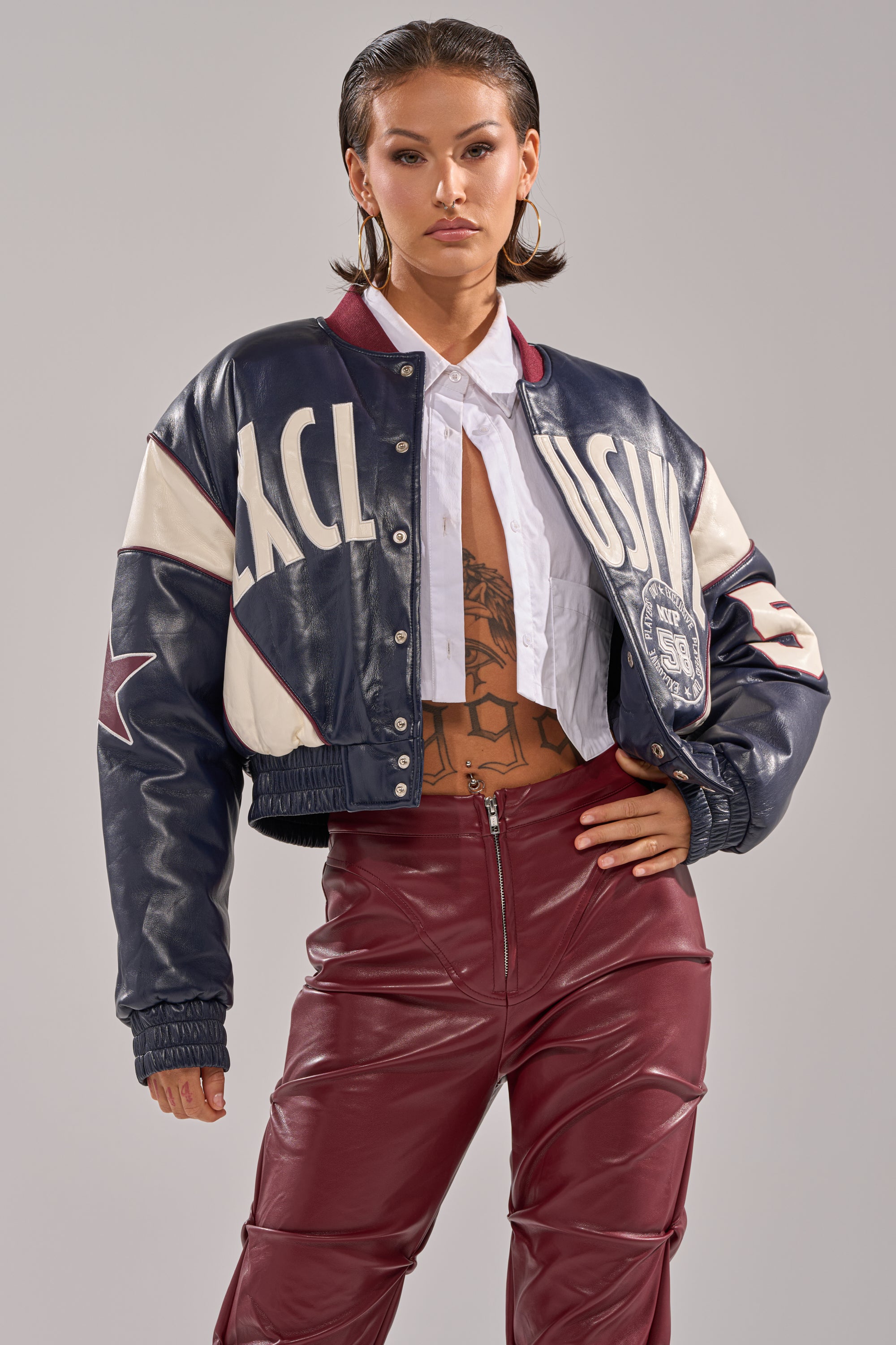 Against a neutral background, a person with slicked-back hair wears the BUTTER LUXE VARSITY BOMBER over an open white shirt, revealing a chest tattoo, paired with high-waisted maroon leather pants.