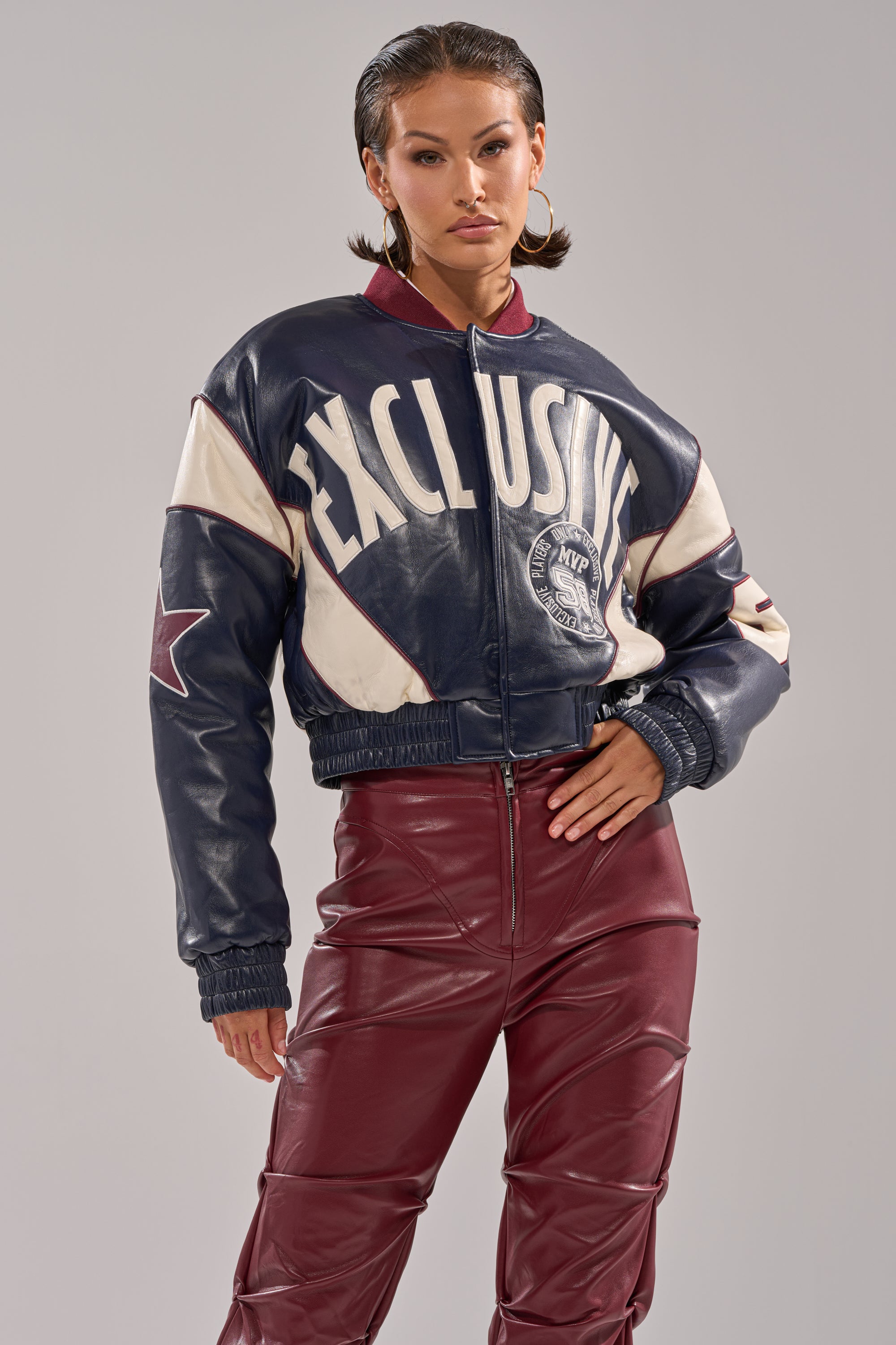A woman poses confidently in the BUTTER LUXE VARSITY BOMBER and high-waisted maroon leather pants against a plain light gray background.