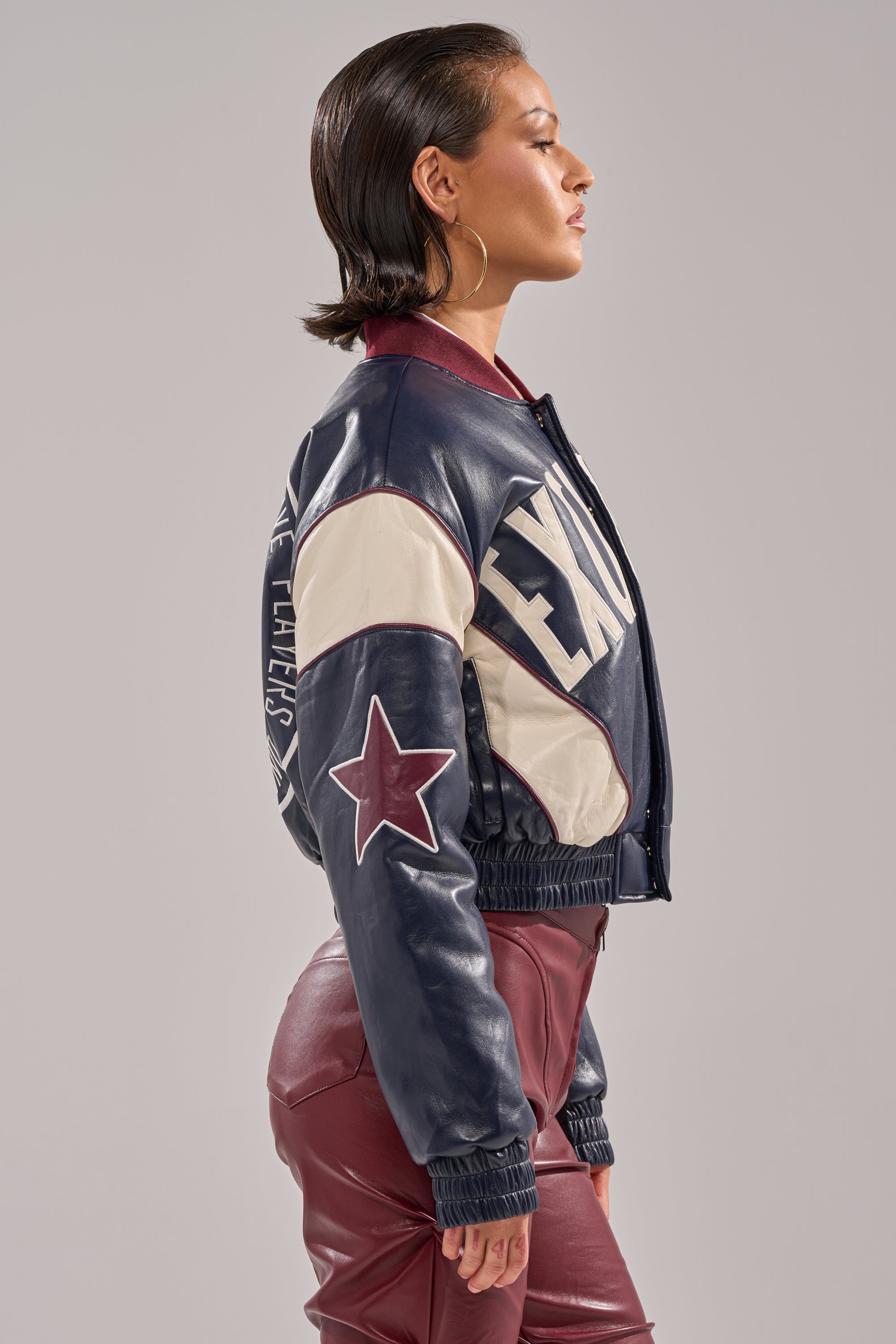A woman in profile wears the BUTTER LUXE VARSITY BOMBER with "TEXAS" on the side, a star on the sleeve, and matching burgundy pants. Her slicked-back hair and large hoop earrings complete her bold, athletic style.