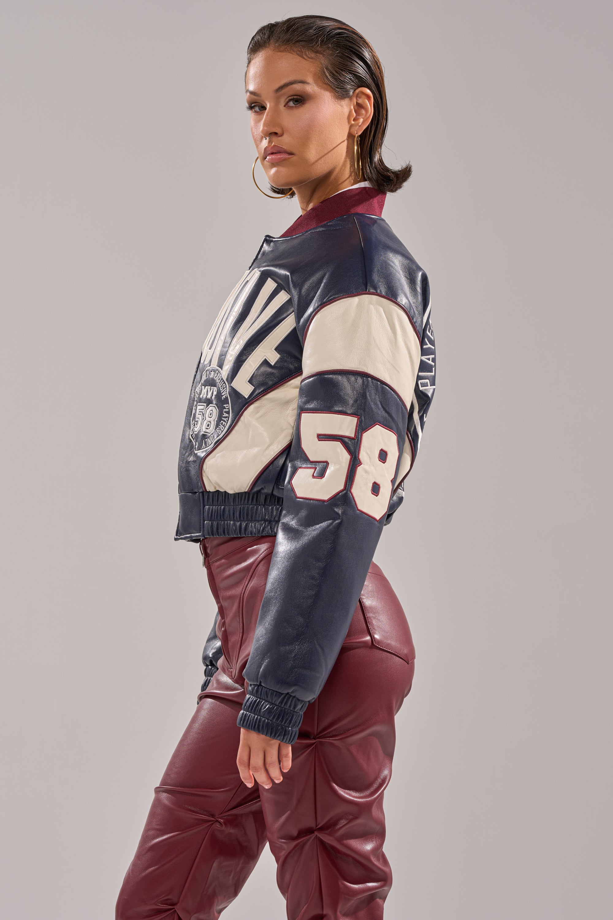 A woman with slicked-back hair confidently poses in the BUTTER LUXE VARSITY BOMBER with "58" on the sleeve and burgundy leather pants, set against a plain gray background.