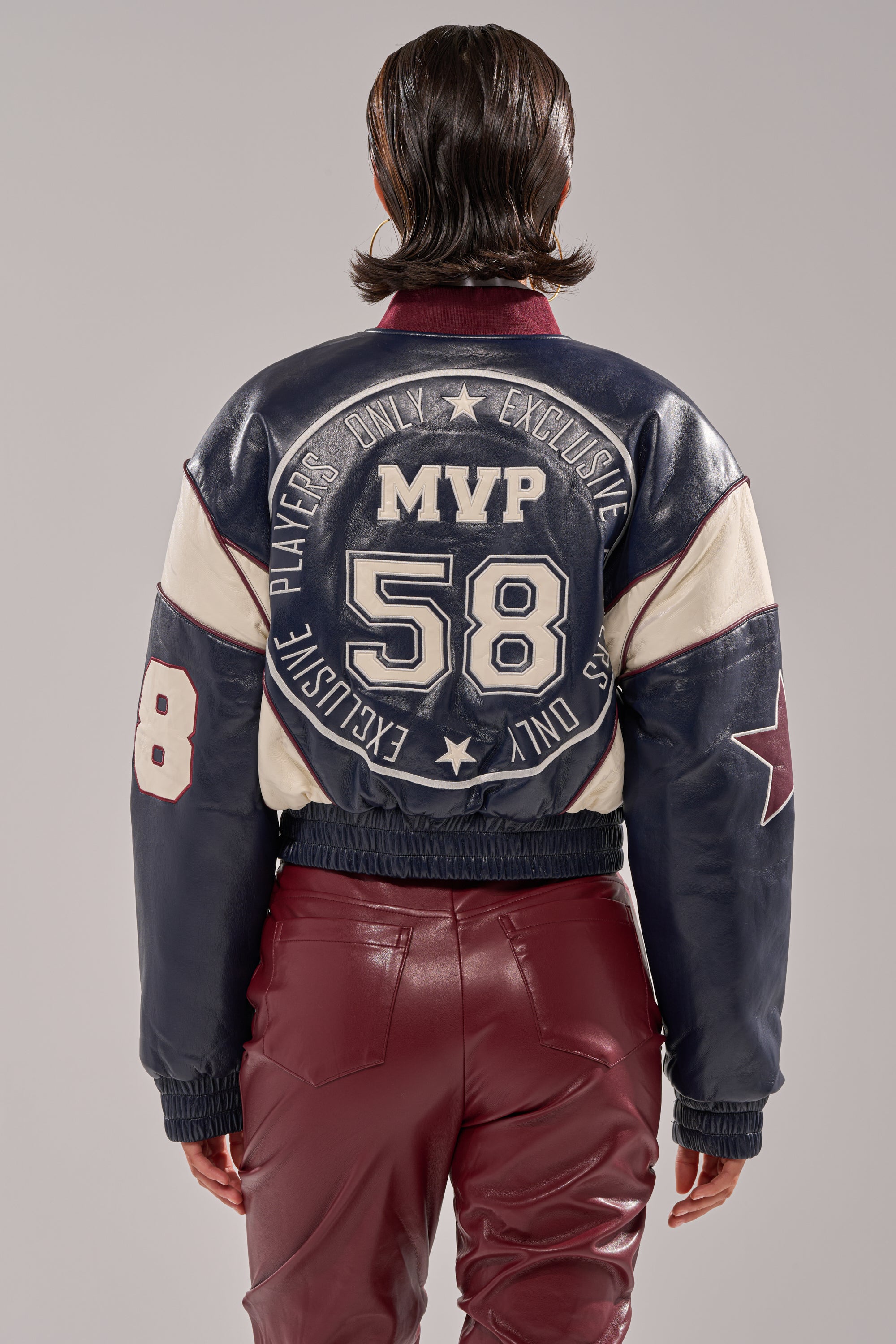 A person stands with their back to the camera, wearing a BUTTER LUXE VARSITY BOMBER in dark blue and cream featuring "MVP 58" and "Players Only Exclusive" on the back, paired with burgundy pants.