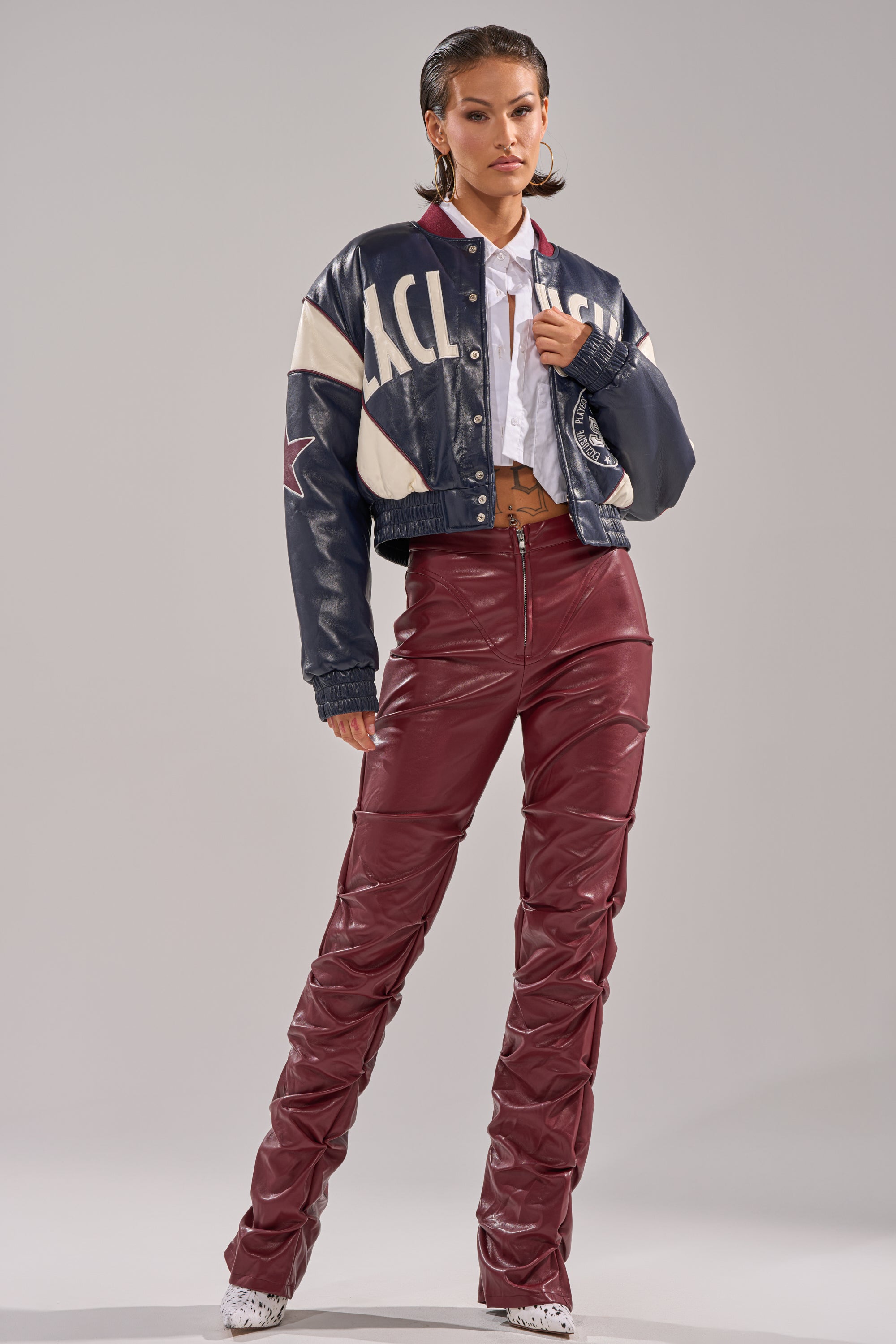 A woman poses confidently against a gray backdrop wearing the BUTTER LUXE VARSITY BOMBER jacket, a white top, high-waisted maroon ruched leather pants, and white shoes.