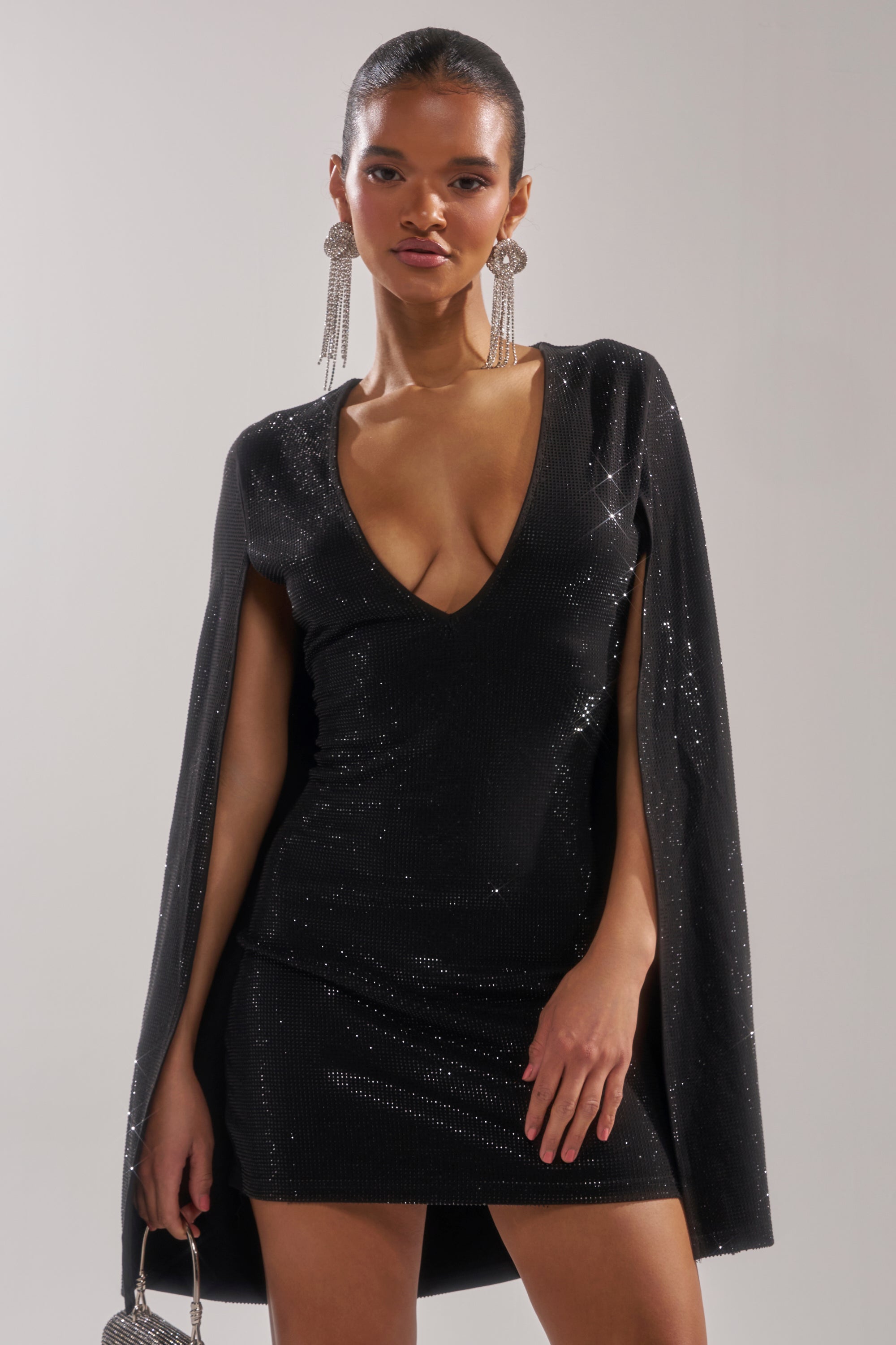 A woman in the LETS DANCE RHINESTONE MINI DRESS, featuring sparkling black rhinestones and cape sleeves, accessorized with long statement earrings and a small silver purse, stands against a plain light background.