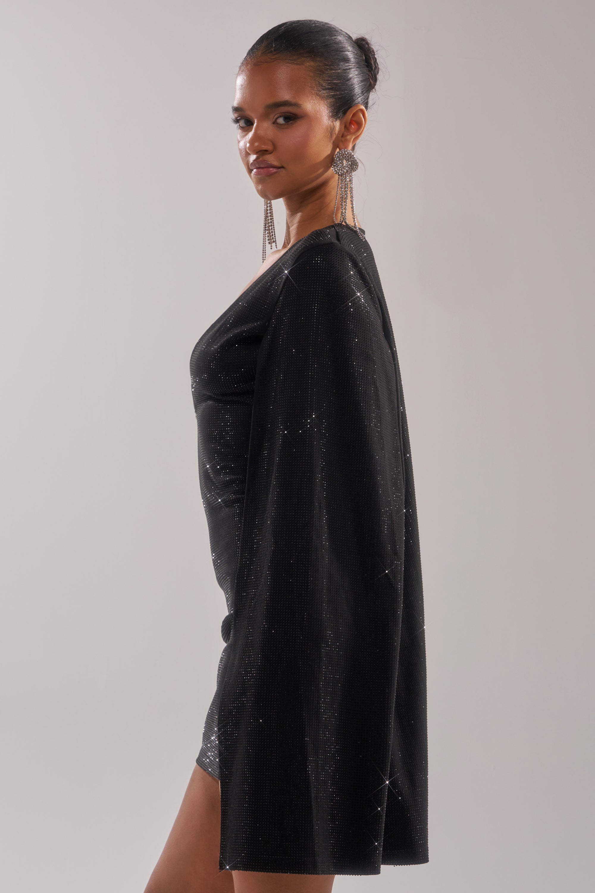A woman with her hair in a bun models the LETS DANCE RHINESTONE MINI DRESS, featuring a sparkly cape-style silhouette and long, dangling earrings as she poses sideways against a plain light gray background.